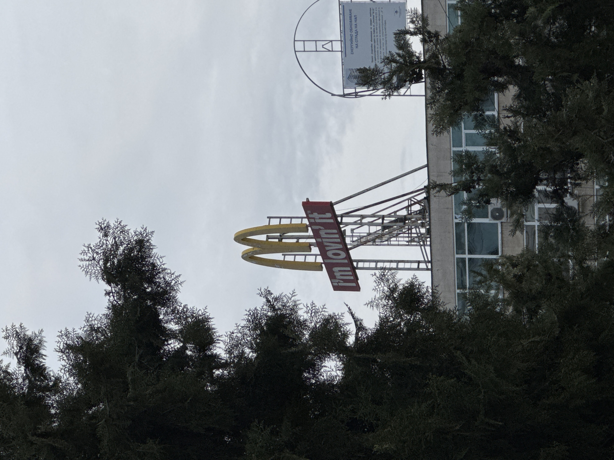 The image depicts a McDonald's restaurant in Sofia, Bulgaria. The golden arches logo and the "I'm lovin' it" slogan are visible on the building's exterior. The building is a multi-story structure with a sign on the roof. Trees frame the foreground. The sky is overcast.