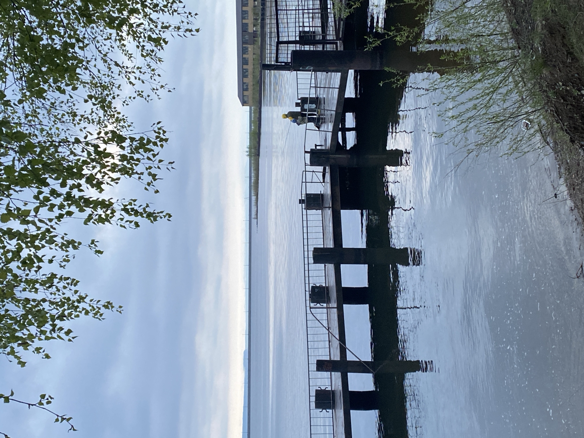 The image depicts a view of the Kama River in the city of Chaikovsky, Russia. A pier extends into the water, constructed of dark wooden posts and metal railings. Two individuals are visible on the pier, both wearing yellow hard hats; one appears to be fishing. A multi-story building is situated on the shoreline near the pier. The sky is overcast with clouds, and trees are visible along the riverbank. The water's surface is rippled, reflecting the sky and pier structure.