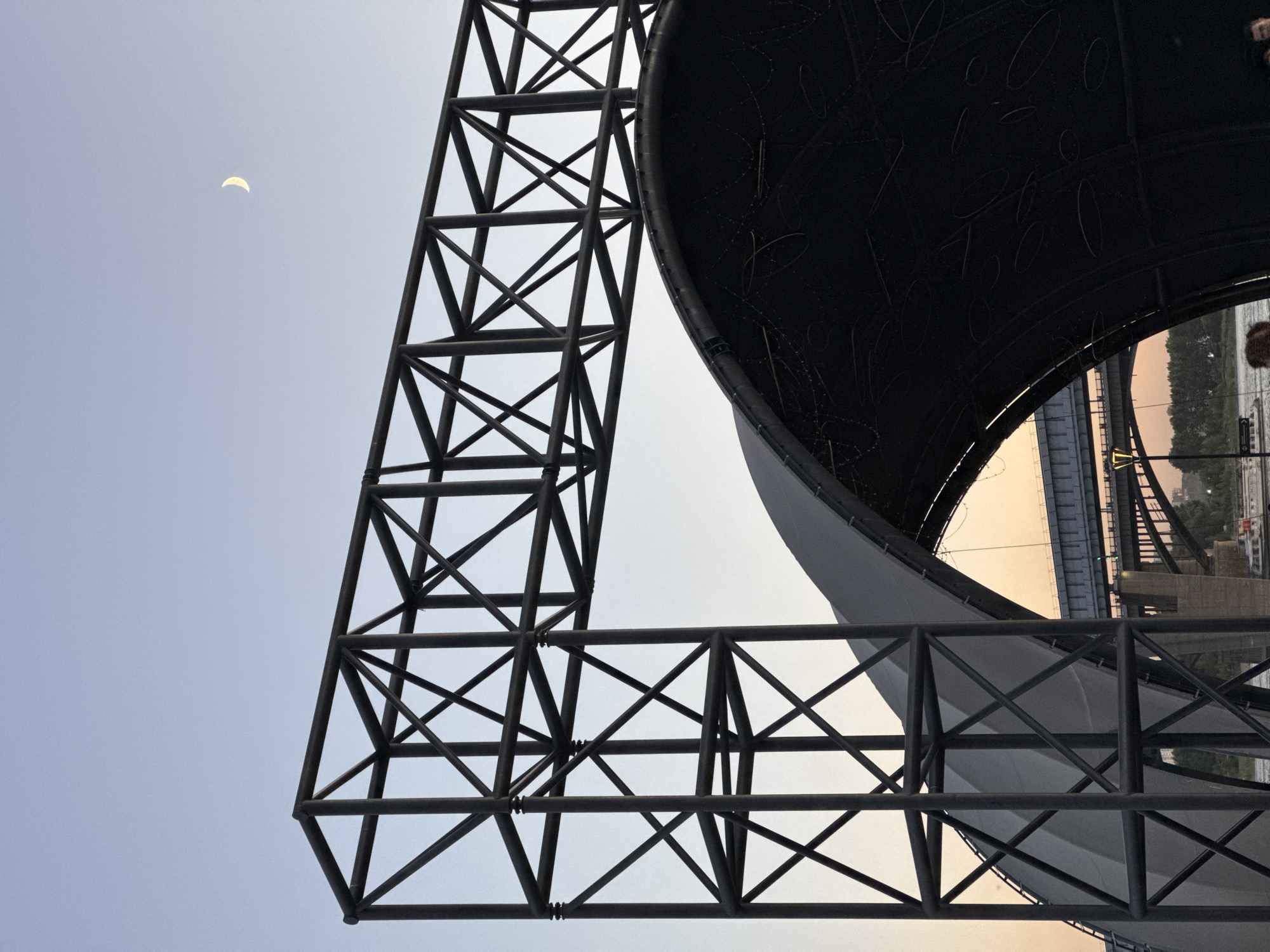 The image displays a low-angle view of a large, dark, curved structure, possibly a dome or canopy, supported by dark metal scaffolding. The scaffolding is composed of interconnected beams forming geometric patterns, creating a sense of industrial architecture against a pale blue sky. In the upper left quadrant of the sky, a crescent moon is visible. Through an opening in the structure and scaffolding, a bridge and a body of water are visible in the background, suggesting an outdoor setting. The sky has a soft, diffused light, indicating it might be early morning or late afternoon. The overall mood is one of scale and structural complexity. There is no visible text in the image. The context provided places this scene in Novosibirsk, Russia.