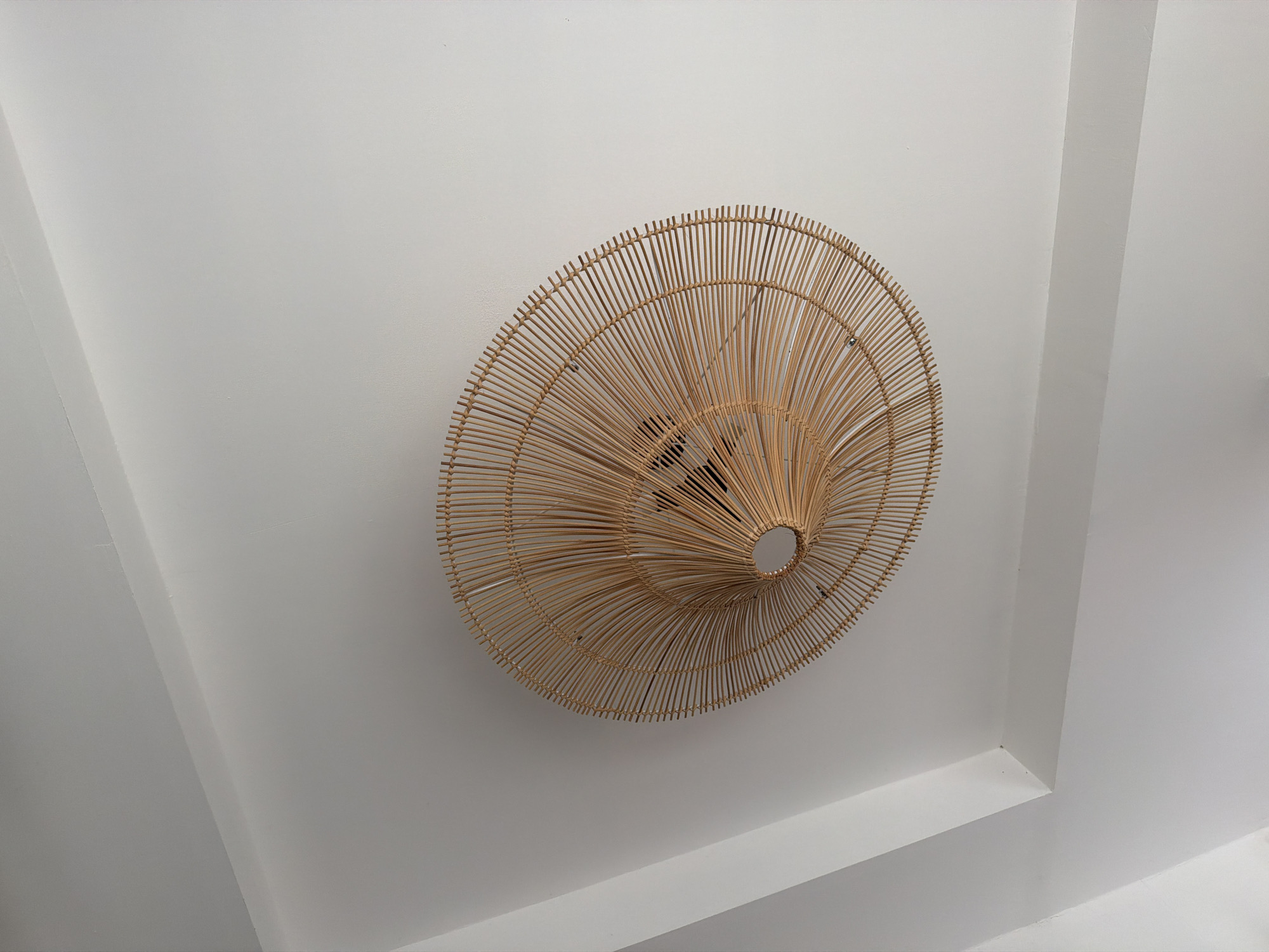 The image shows a close-up, angled view of a natural woven light fixture hanging from a white ceiling. The light fixture is circular and resembles a large, flattened basket or a sunburst design, made of thin, light-colored wooden or bamboo strips. The strips radiate outward from a central opening. The ceiling appears to be a standard residential or commercial ceiling with some architectural angles or recessed lighting details visible in the upper left quadrant of the image. The lighting is neutral, and there are no people or other objects visible in the frame. The scene is indoor and appears to be in a well-lit space. There is no specific visual information in the image to determine the location of Baan Plai Laem, Thailand.