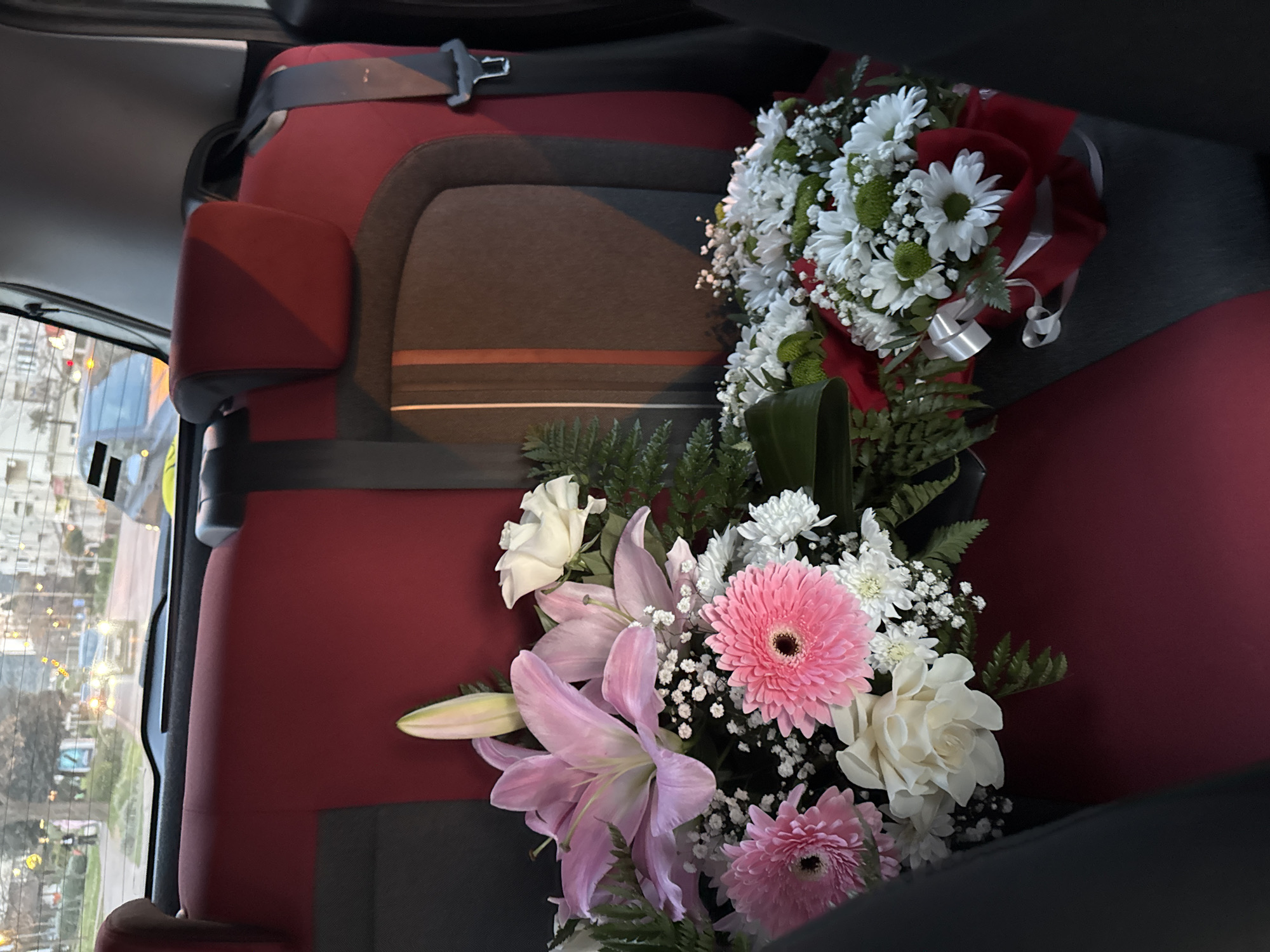 The image depicts the interior of a vehicle, specifically the back seat, which is upholstered in red and grey fabric. Two bouquets of flowers are positioned on the seat. One bouquet contains pink lilies and gerbera daisies, white roses, and white chrysanthemums with greenery. The second bouquet features white daisies, white chrysanthemums, and green accent flowers, tied with red and white ribbons. A safety belt is visible across the seat. Through the window, a cityscape is seen, suggesting the image was taken in Bar, Montenegro. The view shows buildings, streetlights, and vehicles, hinting at an urban setting.