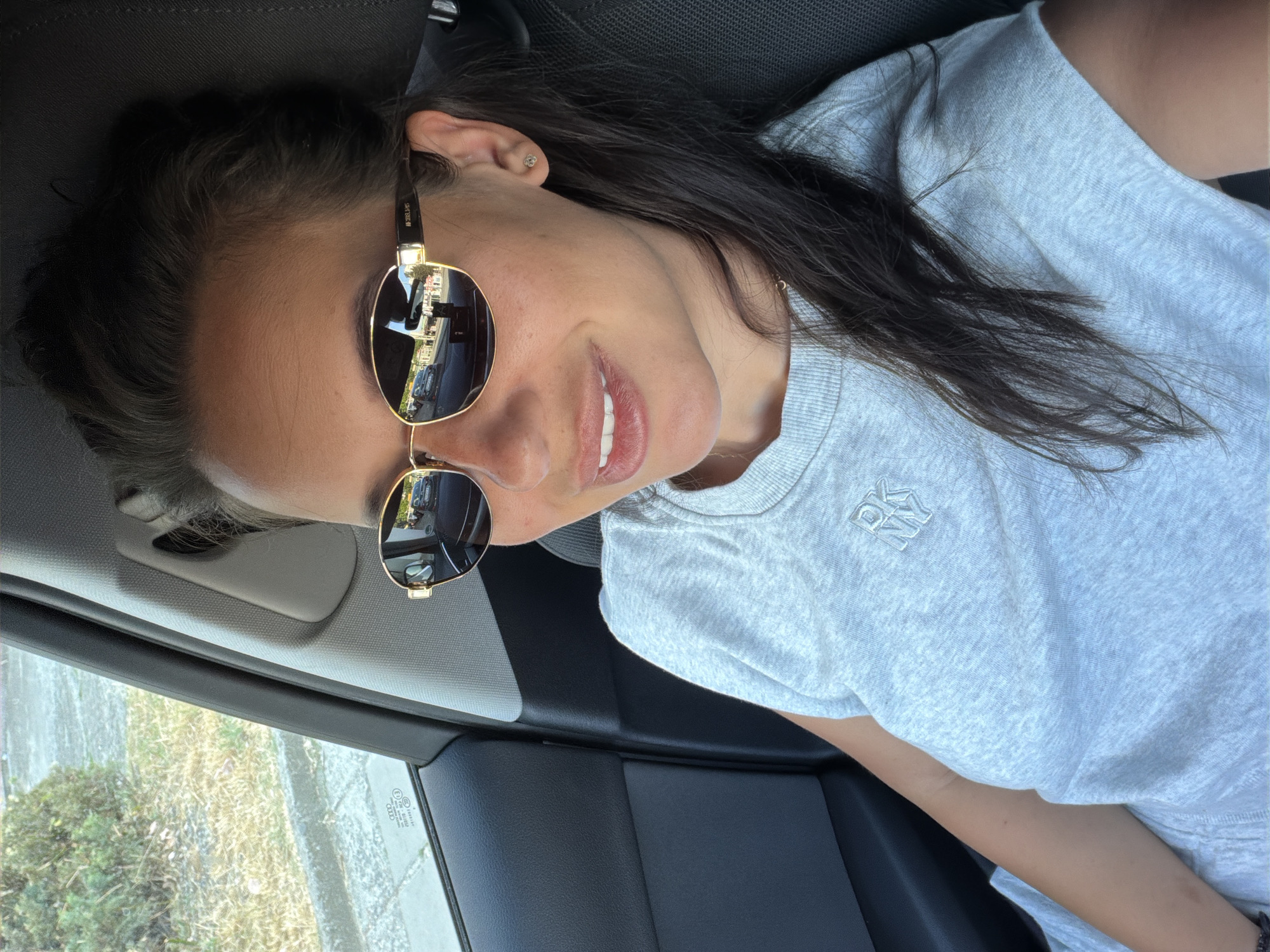 A woman wearing sunglasses and a light grey t-shirt with an embroidered logo sits in the passenger seat of a car. She has long dark hair and is smiling. The interior of the car is visible, including a black leather seat and a grey headrest. Outside the car window, greenery and a street are visible. The woman is likely in Burgas, Bulgaria, given the information provided in the prompt.