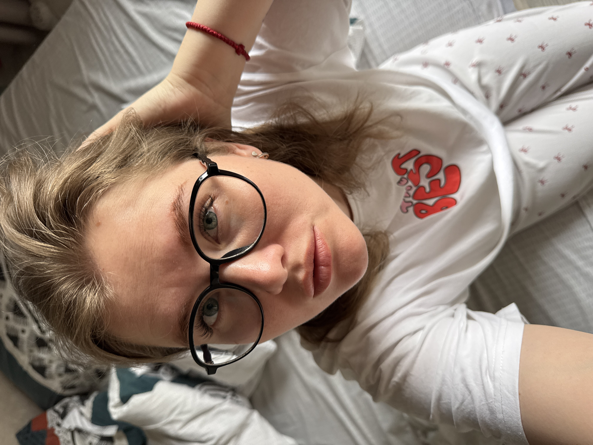 A young woman lies on a gray bed. She wears round black glasses, a white t-shirt with a red and pink print, and white pajama pants with a small red print. Her left hand is raised behind her head, where a red string bracelet is visible. The environment appears to be a bedroom, with a white blanket beneath her. The image gives no indication of the location being Gornyi (Dvurechenskoe S/P), Russia.
