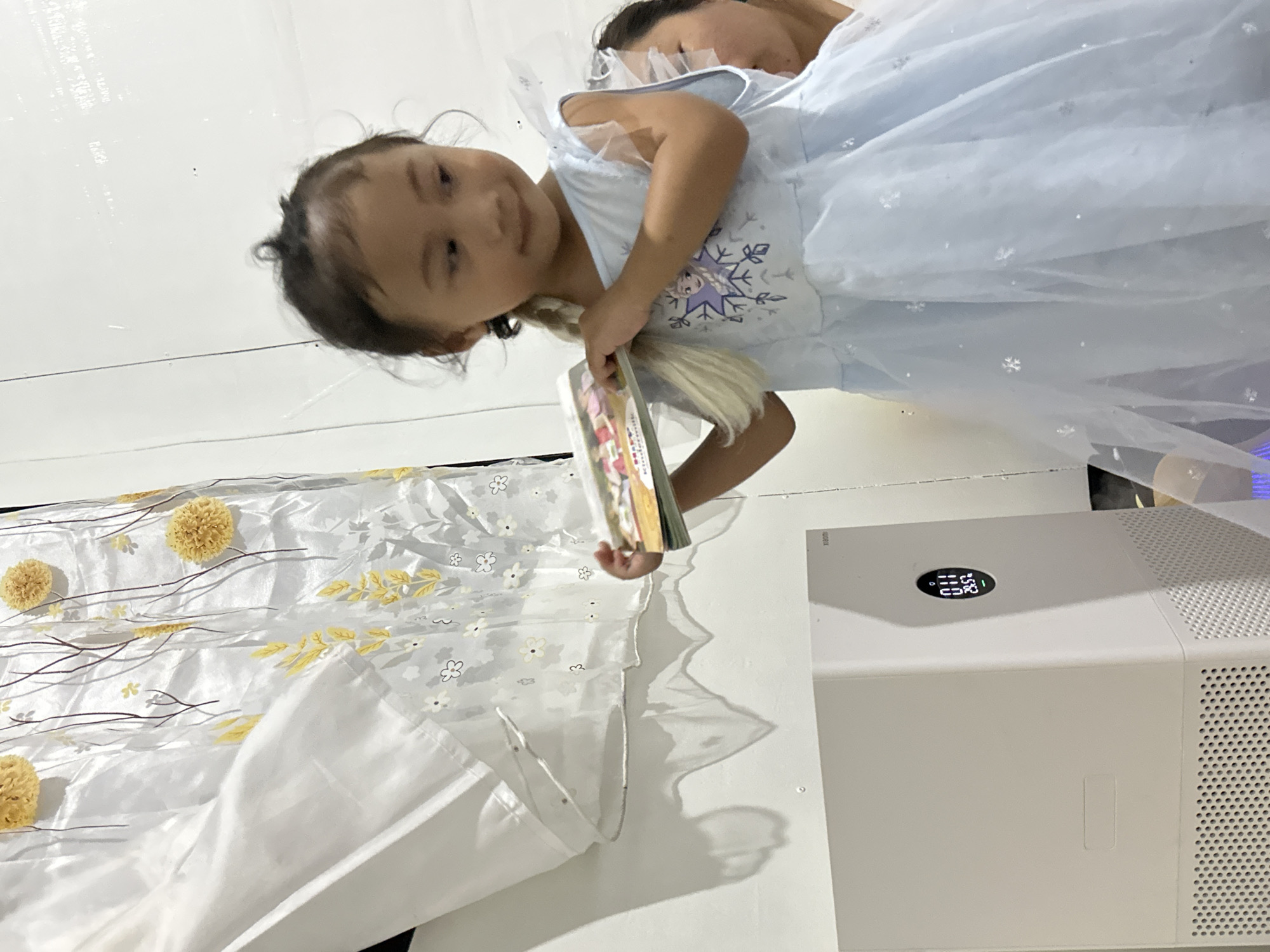The image shows a young girl dressed in a light blue, glittery princess dress with a snowflake design, holding a book. She has dark hair styled in pigtails and is looking towards the camera with a gentle smile. Another person, likely a child, is partially visible behind her, also wearing a similar light blue dress. They are indoors, with a white wall in the background. To the left of the girl, there is a white fabric with a floral print, featuring yellow pom-pom-like flowers and grey branches. To the right, a white rectangular air purifier is visible, displaying a digital screen with some numbers and symbols. The overall setting appears to be a casual indoor environment, possibly a home. The lighting suggests it's daytime. There is no visible text of significance in the image.