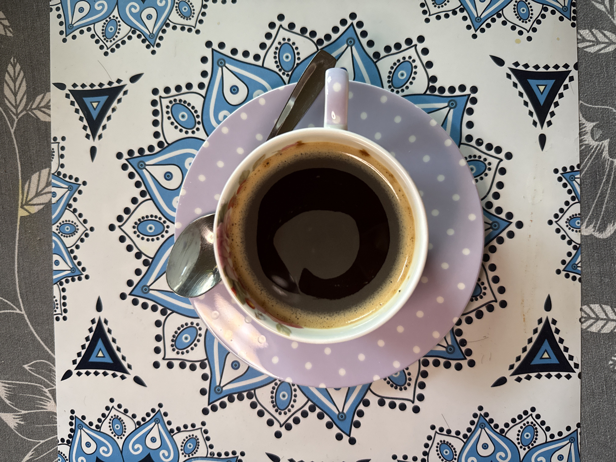 The image presents an overhead view of a cup of black coffee, centrally placed on a patterned surface. The coffee cup has a white interior and is filled with dark coffee, showing a thin layer of crema on the surface. It rests on a light purple saucer adorned with white polka dots. A metallic spoon is positioned within the cup, with its handle extending onto the saucer.
The cup and saucer are situated on a white surface that features an intricate, repeating pattern of blue and black mandala-like designs. These designs include stylized teardrop or floral shapes with "eye" motifs, surrounded by small black dots, as well as distinct triangular elements in black and blue. This patterned surface appears to be a placemat or tray. To the left and right edges of the frame, a grey fabric with a subtle pattern of light-colored leaves or foliage is visible, likely serving as a tablecloth or another surface beside the placemat.
The scene depicts a simple, quiet moment, suggesting a coffee break in an indoor setting. No individuals are present, and there is no visible text. The time of day and external weather conditions are not discernible from the image. The location is contextualized as potentially being in Pleven, Bulgaria.