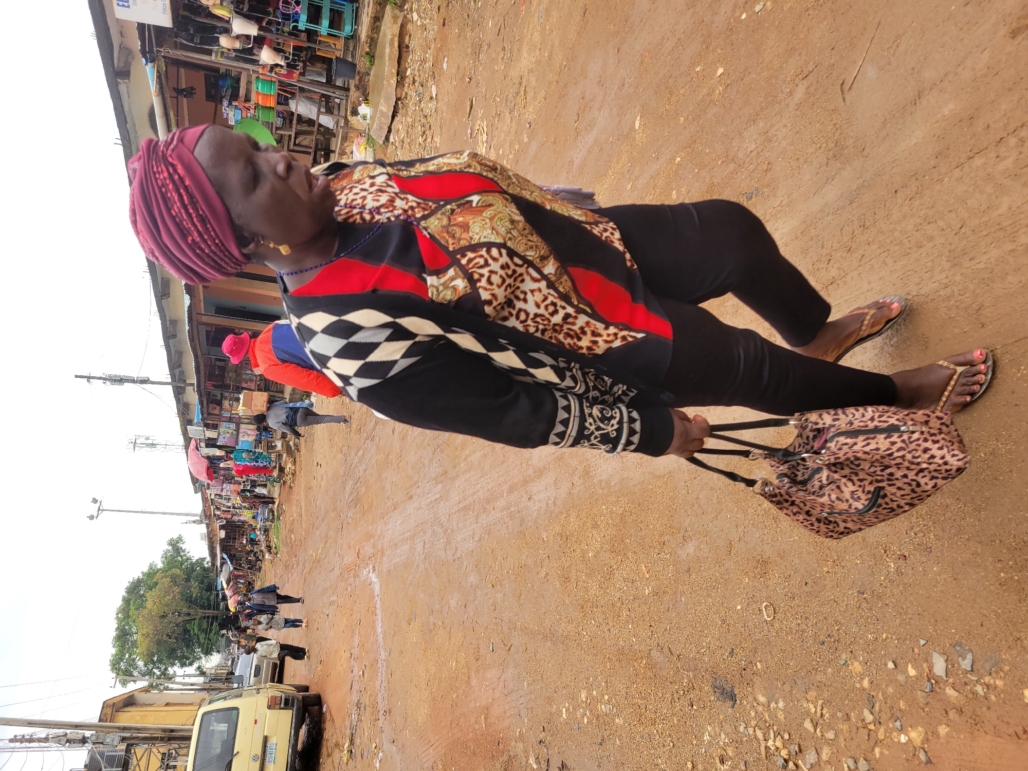 A woman is depicted walking along a road in Jos, Nigeria. She has dark skin, wears a red headscarf, a patterned shirt, black pants, and brown sandals. She carries a leopard print handbag. The road is dirt and appears to be in a market area. People and vehicles are visible in the background.