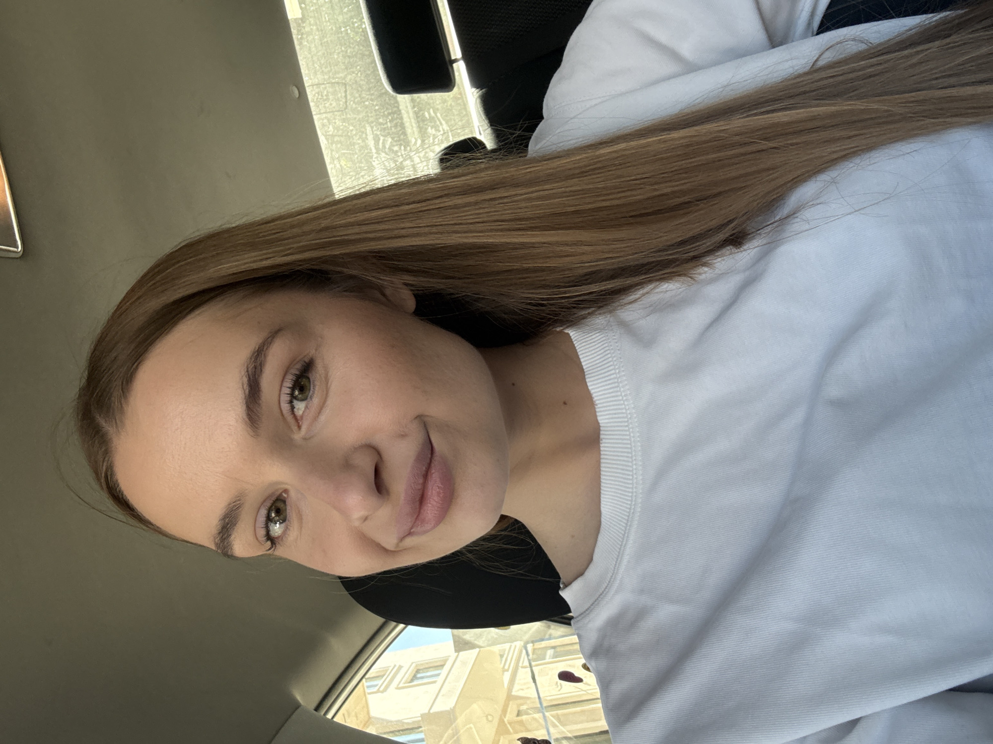 A woman is in a car. She has long, straight, brown hair and green eyes. She is wearing a white sweater. The car appears to be parked, with a building visible through the windshield. Based on the architectural style visible through the windshield, it is likely the location is in Budva, Montenegro, though this cannot be definitively determined from the image alone. The presence of the car suggests transportation as an activity. There is no clear interaction in the image, as it appears to be a self-portrait. The environment includes the interior of a car, and an urban exterior scene.