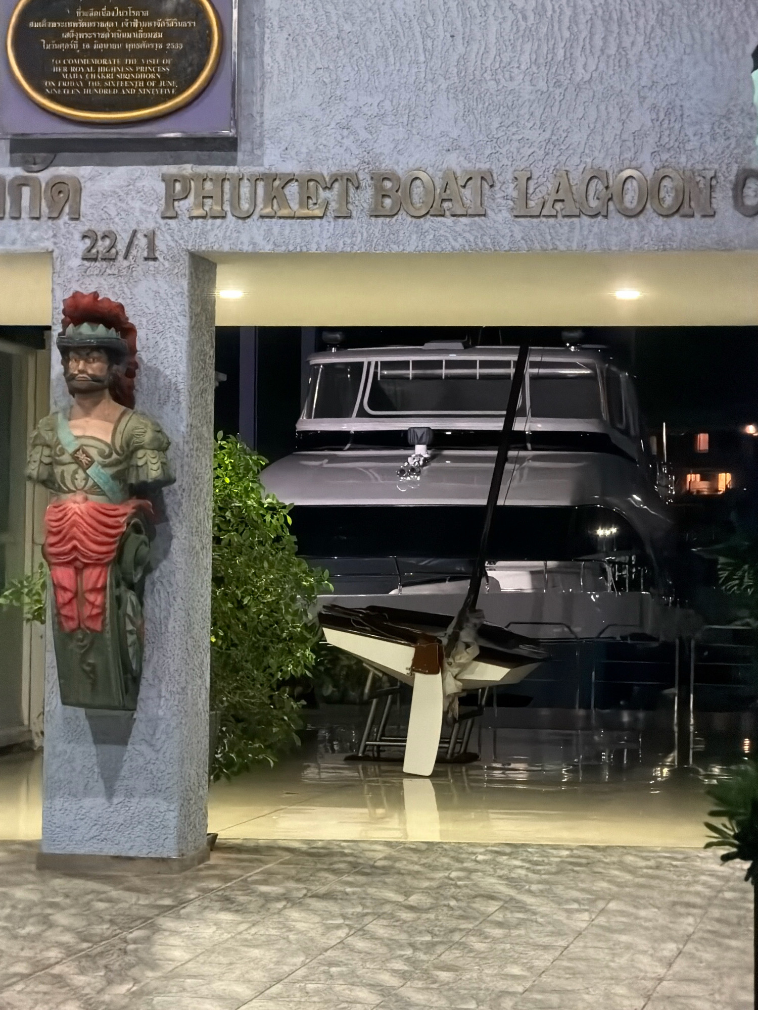 The image captures a boat jammed into the entrance of the Phuket Boat Lagoon in Ko Kaeo, Thailand. A statue of a man in historical attire stands to the left of the entrance, partially obscuring the view of the interior. The boat is a large, modern vessel with a white hull and black trim, its bow protruding into the building. The entrance itself features large, metallic lettering that reads "PHUKET BOAT LAGOON". The ground appears to be flooded, reflecting the light from the interior and the boat. The overall scene suggests a recent accident or an unusual placement of the boat within the facility.