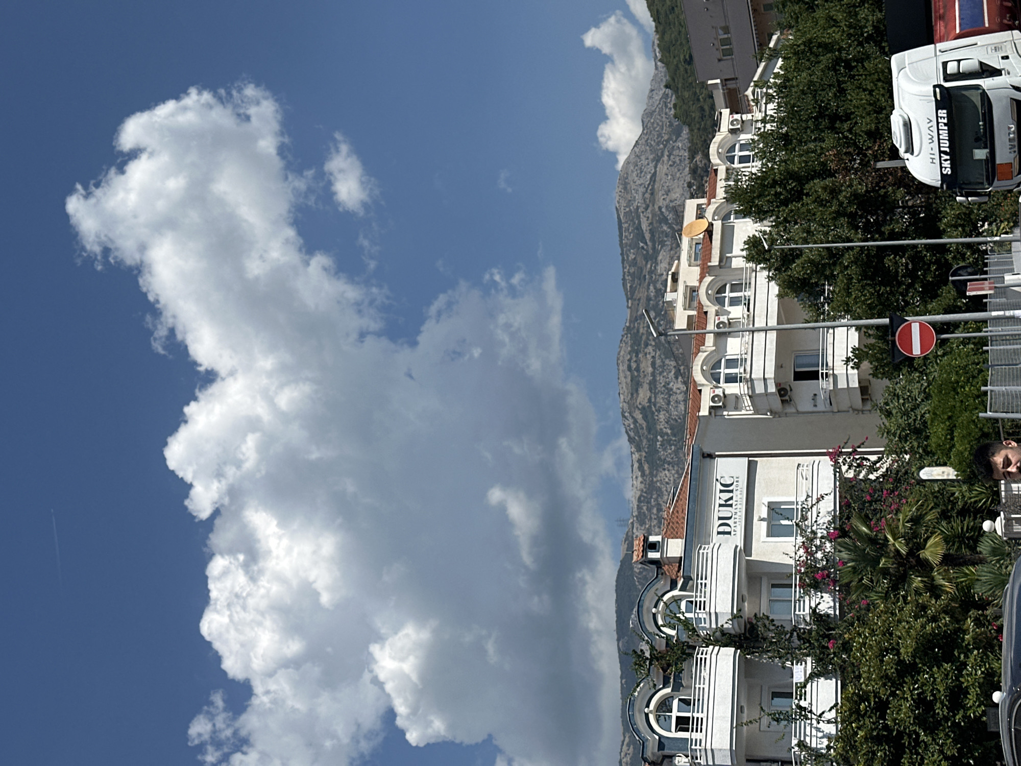 The image shows a bright, sunny day in Budva, Montenegro, with a clear blue sky punctuated by large, puffy white clouds. In the foreground, a white truck with "HI-WAY SKY JUMPER" written on its side is partially visible on the right. Adjacent to the truck, a traffic sign with a red circle and a horizontal white bar indicates "no entry." Behind this, a modern, white apartment building with arched windows and balconies is visible. A sign on the building reads "DUKIĆ APARTMANI - SOBE." Lush green trees and flowering vines with pink blossoms partially obscure the lower floors of the building. In the background, a range of rugged, treeless mountains forms the horizon. A faint contrail from an airplane is visible in the upper portion of the sky.
