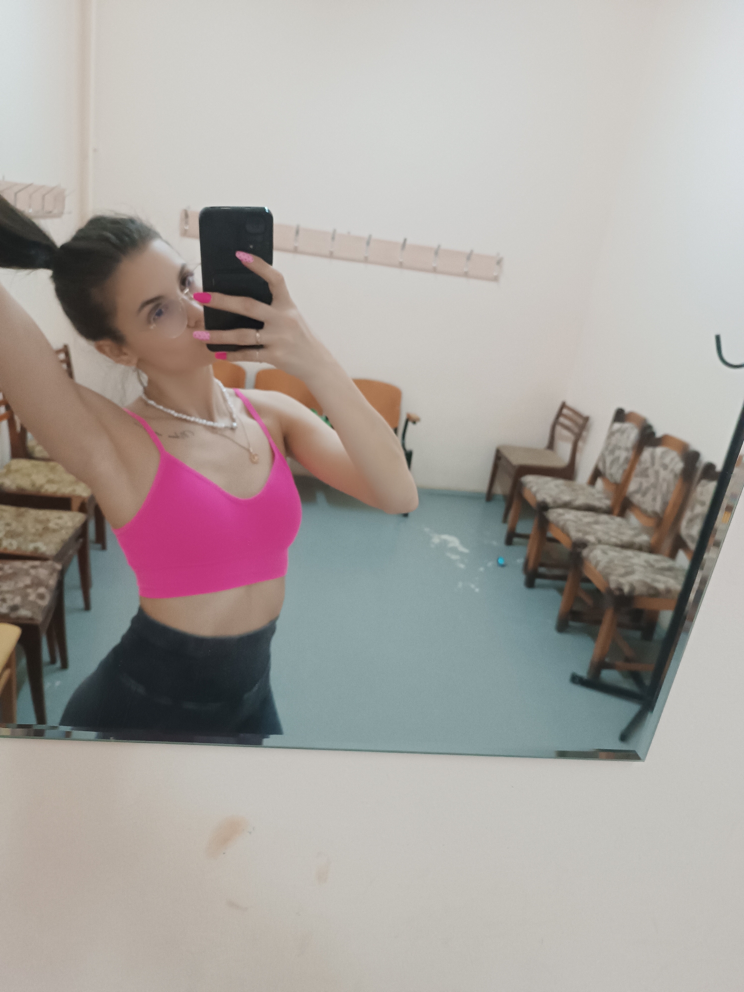 A person is taking a mirror selfie in what appears to be a waiting room. The individual has brown hair pulled back in a ponytail, is wearing a pink sports bra and dark pants, and is holding a black smartphone. They are wearing a pearl necklace and eyeglasses. The waiting room contains several wooden chairs with patterned upholstery, coat hooks along the wall, and a blue floor. Streaks of what seems to be dried soap are on the floor. The location is in the city of Unknown, Unknown.