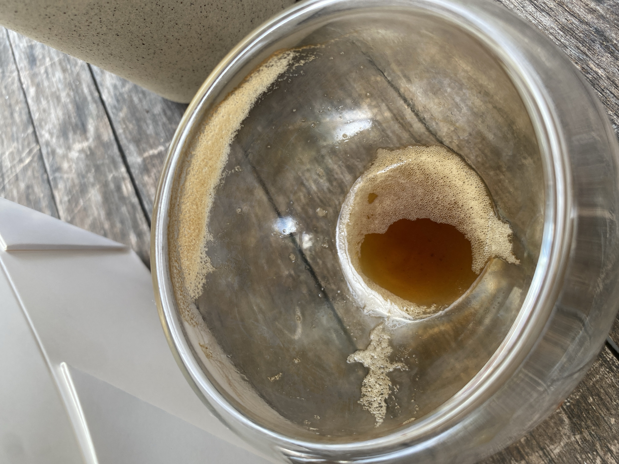 A top-down, medium shot captures the remnants of a beverage in a glass bowl. The bowl, with clear glass, sits on a weathered, grey wooden surface, likely a table. The liquid, a deep amber color, forms a small pool at the bottom of the bowl, surrounded by a foamy residue clinging to the inner glass walls. The residue also forms a thin, light brown ring around the upper edge of the bowl. The background is blurred, but hints of a white rectangular object are visible to the left, and a speckled, beige cylindrical object is partially visible to the right. The overall impression is one of a recently finished drink, likely enjoyed outdoors given the rustic table. The image is clear and well-lit, with the focus on the drink's residue within the glass.