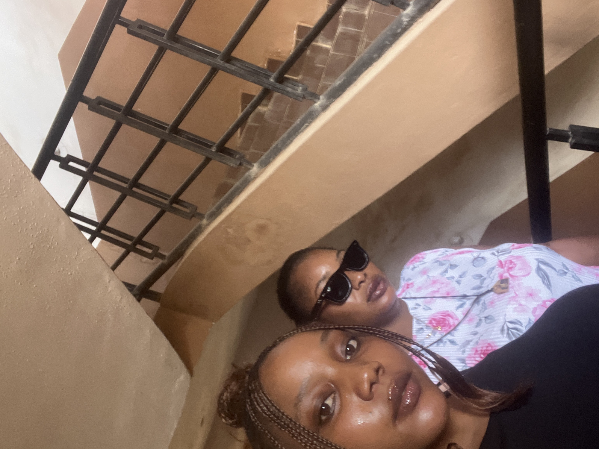 Two young Black women are standing on a staircase with a dark metal railing. The woman in the foreground, on the left, is closer to the camera and has braids in her hair, which are pulled up into a bun. She is wearing a black top and has a shiny lip gloss. The woman behind her, on the right, is wearing sunglasses and a white top with a pink floral pattern. She is also wearing a necklace with a pendant. The staircase is a light peach color with a dark metal railing. The background shows more of the staircase and the wall. The scene appears to be indoors. The image does not contain any specific visual elements that would allow for a precise identification of the location within the city of Jos, Nigeria.