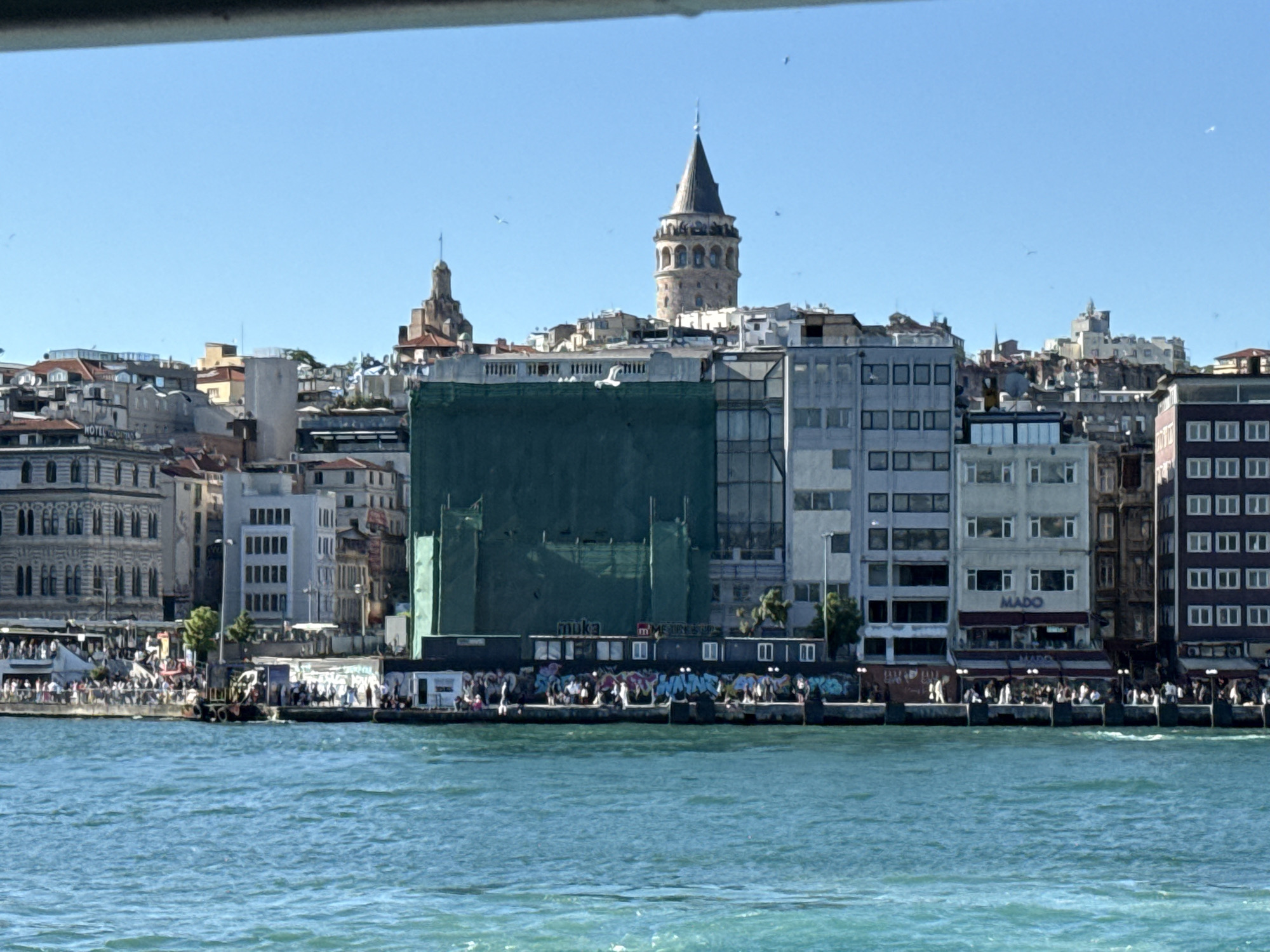 The image captures a waterfront view in Beyoğlu, Turkey, featuring a bustling scene along the shoreline and a backdrop of varied architectural styles. Prominently, the Galata Tower rises above the skyline. A large green construction screen partially obscures a building in the midground. A waterfront promenade is lined with shipping containers covered in graffiti and packed with people. Further along the water's edge, there is a MADO cafe and a number of people seated at tables. The water in the foreground is light blue. The sky is bright and clear with a number of seagulls in the distance.