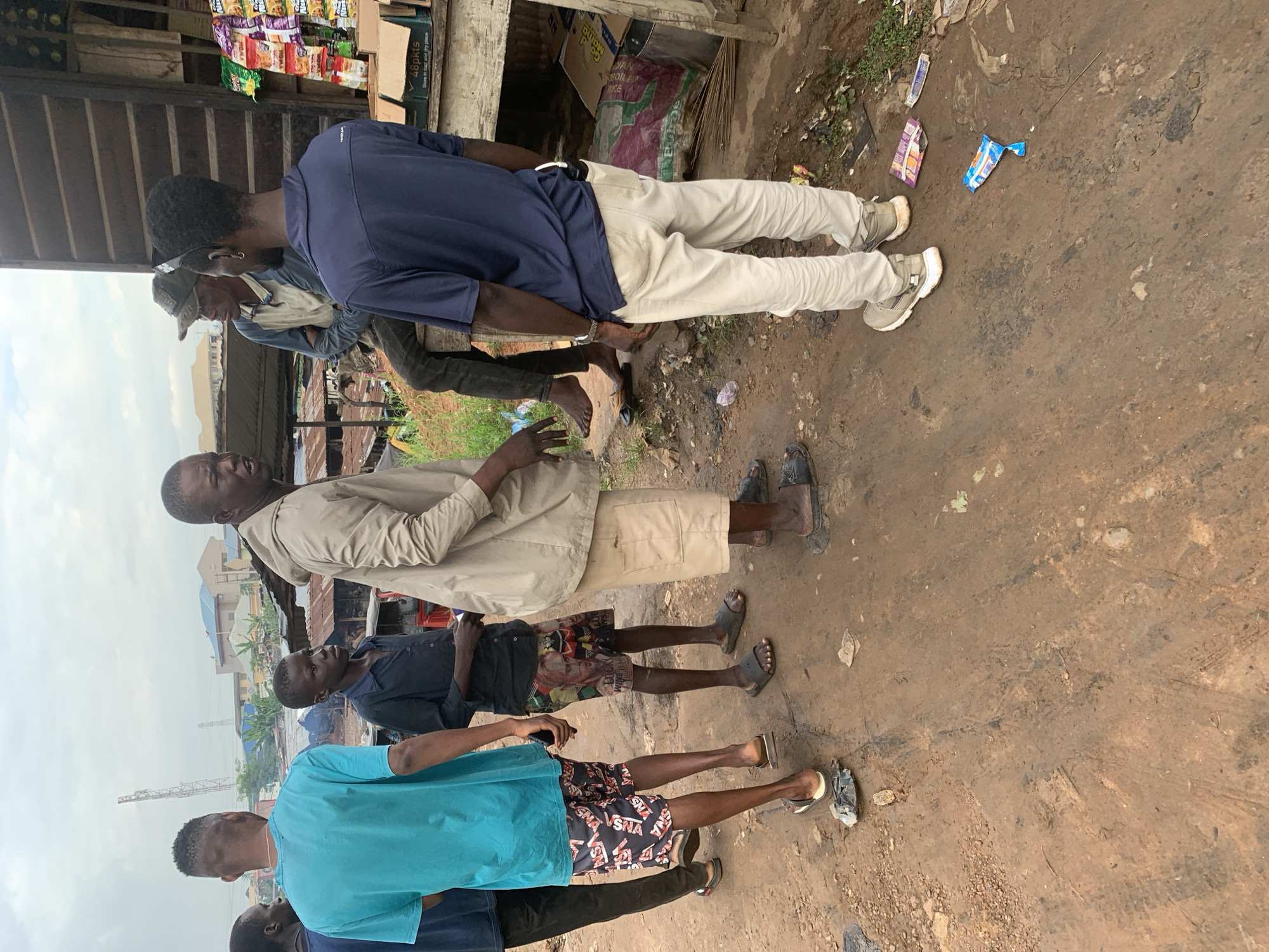The image captures a street scene in Shagamu, Nigeria, with several individuals gathered near a makeshift stall. The ground is unpaved and appears to be dirt and mud, with scattered debris. In the foreground, five men are present. One man, wearing a teal t-shirt and patterned shorts, stands on the left. Next to him, a younger man in a dark blue shirt and patterned shorts is also standing. Further to the right, a man in a light beige jacket and light-colored shorts is speaking with two other men. One of these men wears a dark blue polo shirt and light-colored pants, while the other, seated, is wearing a cap and a light-colored long-sleeved shirt. Behind the group, a stall is set up with various goods displayed, including packaged snacks and beverages. The overall environment suggests an outdoor market or roadside commerce.