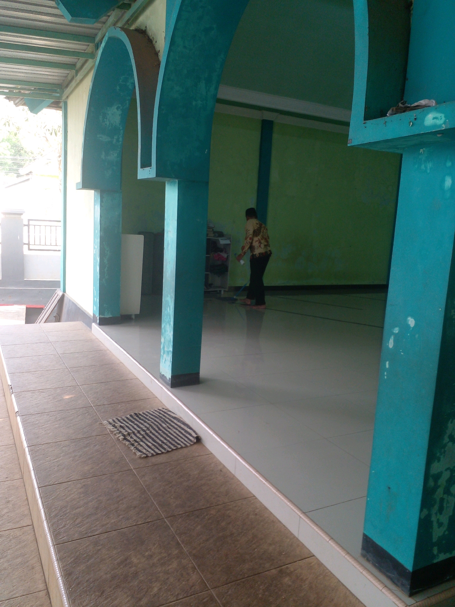 A person is mopping the floor in an indoor space. The person is wearing a patterned shirt and dark pants. The room has light green walls and white tiled floors. There are teal-colored pillars and archways. The space appears to be a public building, possibly a mosque, given the architectural style. The scene is well-lit, with natural light coming from an opening on the left side of the image. The overall impression is one of cleanliness and order.