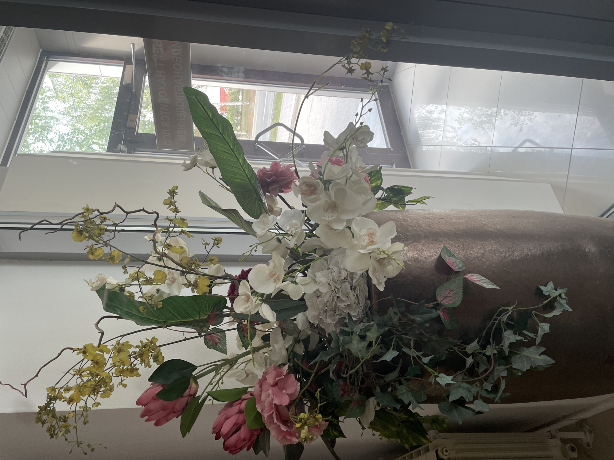 A large, decorative floral arrangement is positioned in a bronze-colored vase next to a glass door in what appears to be an interior space. The arrangement features a variety of flowers, including pink proteas, hydrangeas, white orchids, and small yellow blossoms, along with large green leaves and trailing ivy. The glass door leads to an outdoor area with trees and a street visible. A sign with Cyrillic text is affixed to the glass door, partially obscured by the floral arrangement and the reflection on the glass. The floor inside is tiled. To the left of the arrangement, a section of a white radiator is visible. The lighting suggests it is daytime, with natural light entering from the doorway and possibly from overhead. No people are present in the scene. The overall scene suggests a commercial or residential interior space decorated with a prominent floral display. The sign on the door is likely a business name or informational notice, and the location in Surgut, Russia, is inferred from the Cyrillic text on the sign.
