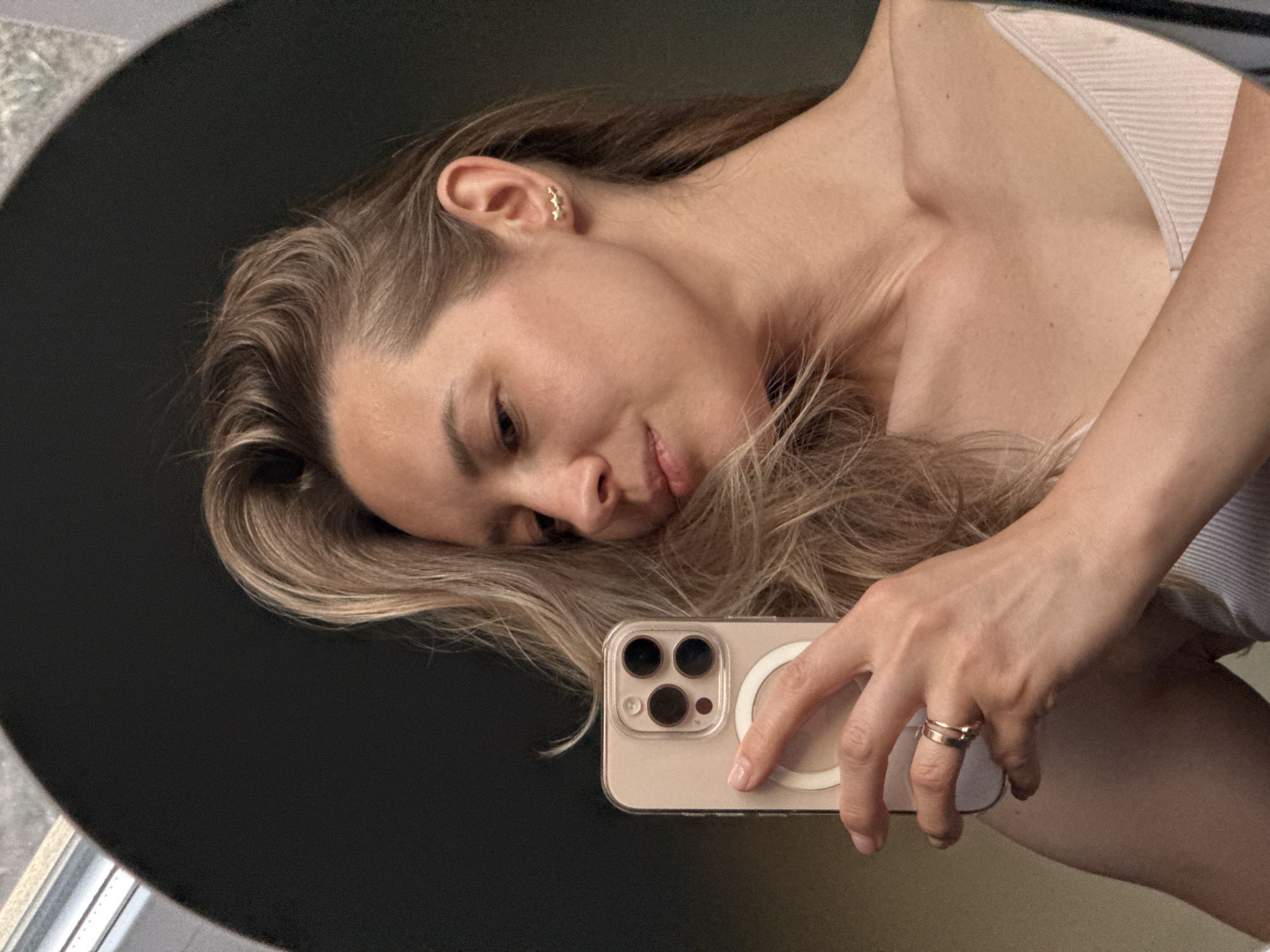 A woman is taking a selfie in a mirror. She is wearing a white top, and her hair is blonde. She is holding an iPhone with a clear case. The mirror is round and has a black background. The location is unspecified in Chaikovsky, Russia.