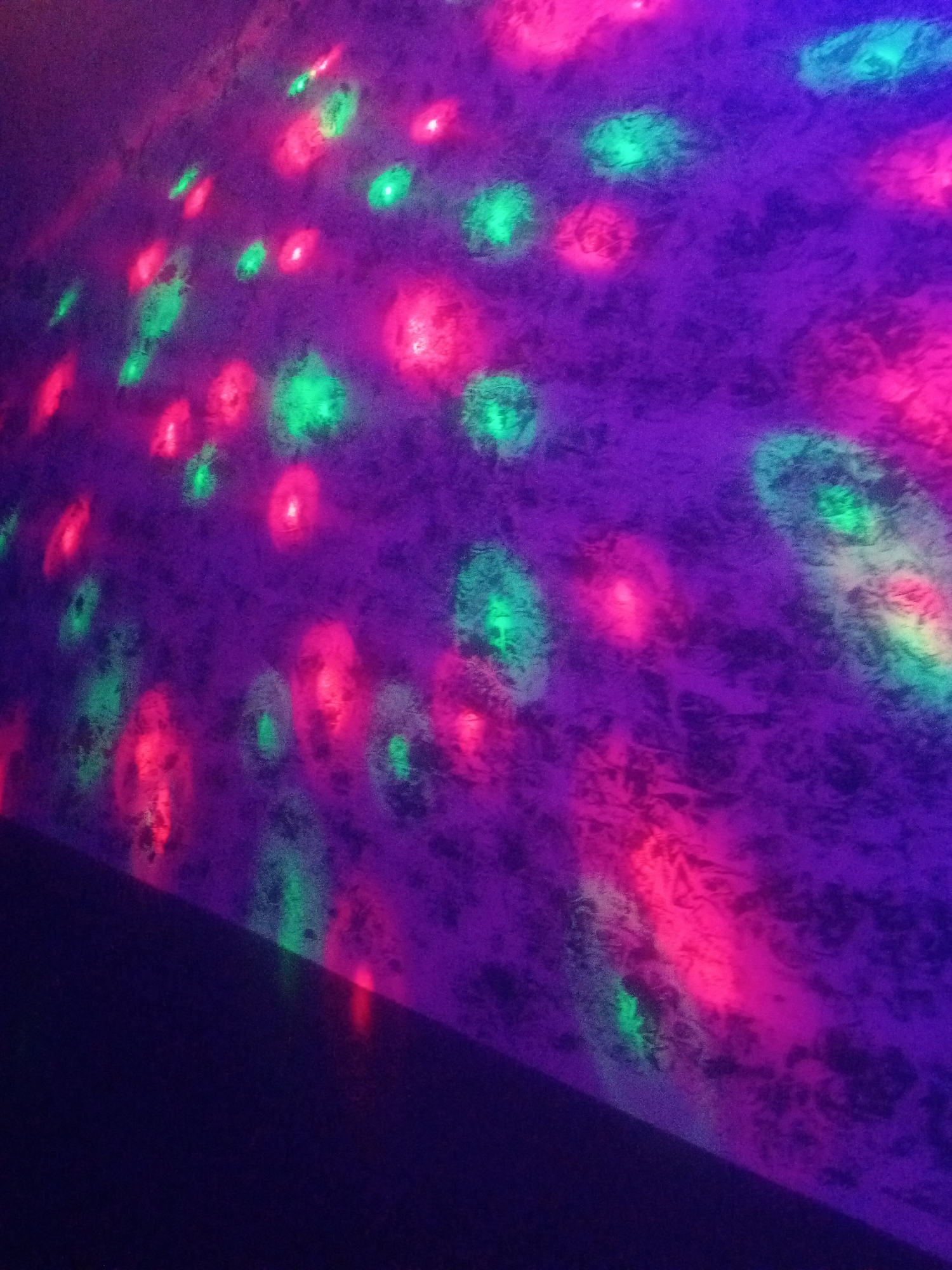 The image depicts a textured, purple wall illuminated by scattered red and green lights. The lights cast irregular, diffused circular and oblong patterns across the wall's surface. Some areas of the wall appear darker, indicating shadow or less direct light. The overall impression is of a dimly lit, atmospheric space, possibly a party or entertainment venue. There are no people or discernible objects visible in the image, and the focus is entirely on the interplay of light and the textured wall. The location of Sabon Gari Nassarawa, Nigeria, is an assumed context and not visually represented in the image itself.