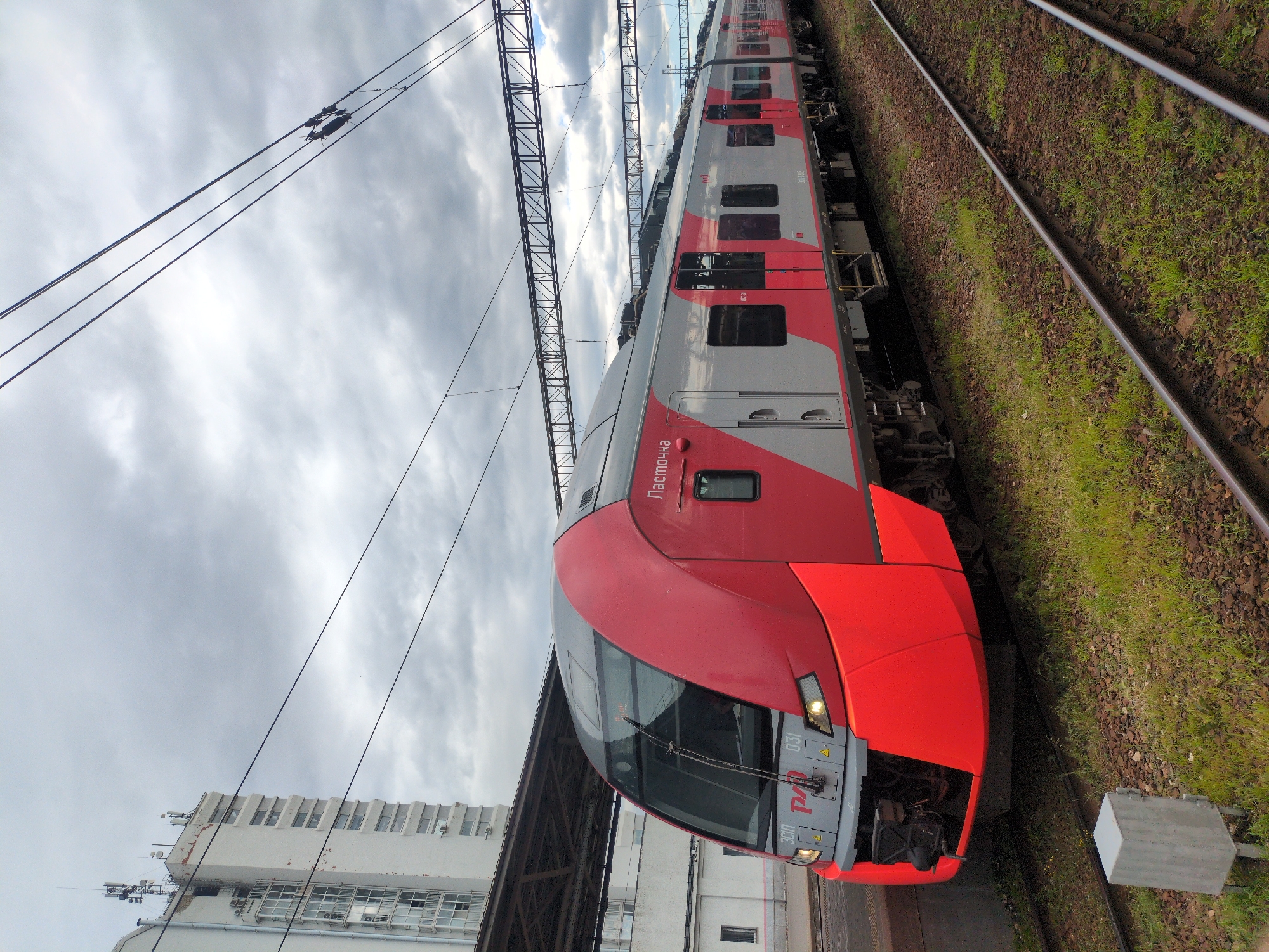 A high-angle, rotating shot captures a red and gray commuter train stopped at a station. The train has a sleek, modern design with large windows and the inscription "Ласточка" on its side. The train is positioned alongside a grassy embankment and a set of railway tracks. Overhead, a network of power lines stretches across a cloudy sky. In the background, a white, multi-story building with many windows is visible. The image is taken during the daytime, under overcast weather conditions. The scene appears to be a railway station in Nizhny Novgorod, Russia.