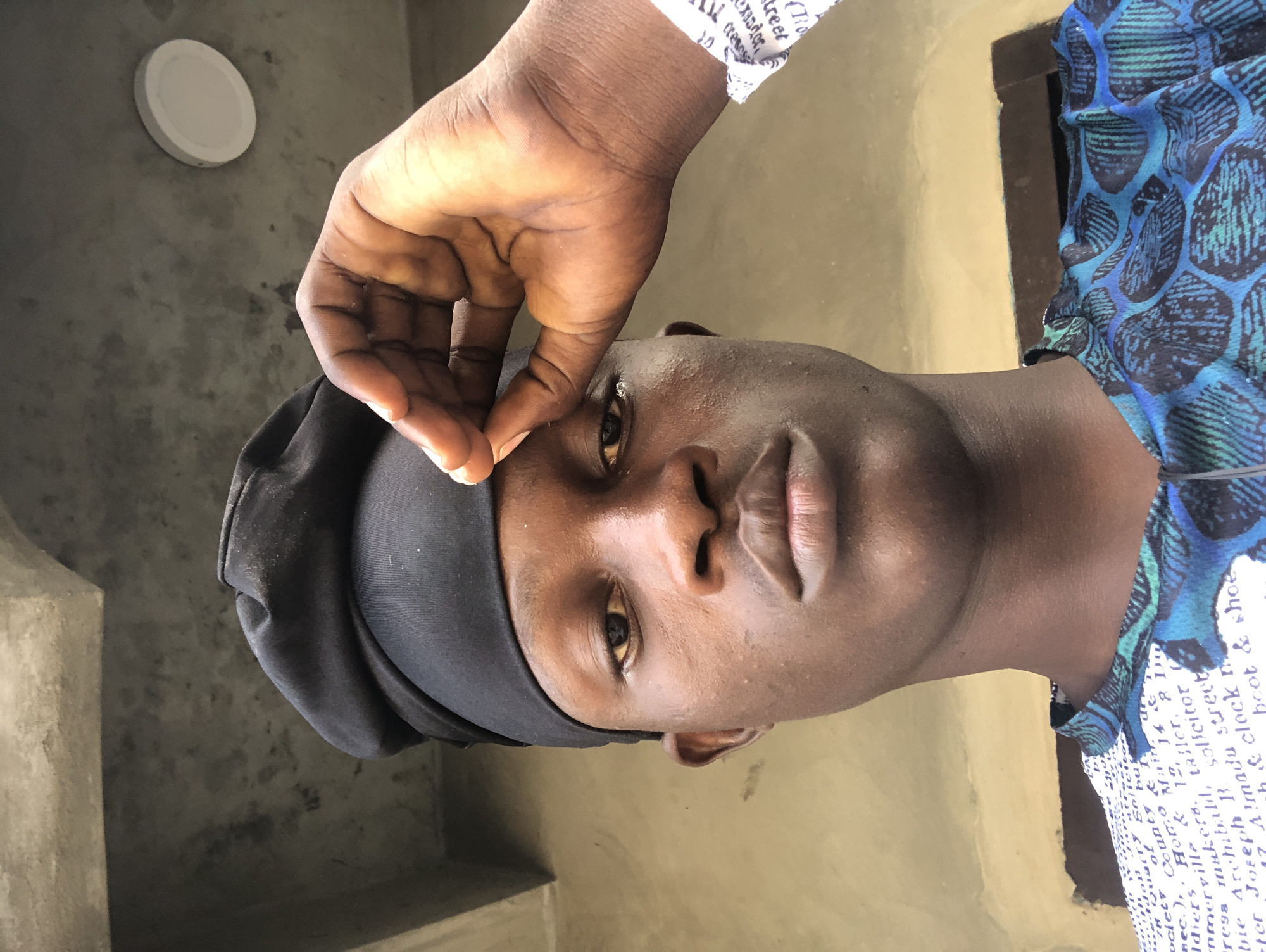 In Ado, Nigeria, a close-up, low-angle selfie captures a young Black male from the chest up, facing the camera. He wears a black headwrap and a patterned shirt with blue and black designs. His right hand is raised, with his index finger and thumb touching the top of his headwrap. The background is an outdoor scene with a concrete wall and ceiling, and a white circular light fixture. Text is visible on his shirt and sleeve, but is not legible. The lighting suggests it is daytime.
