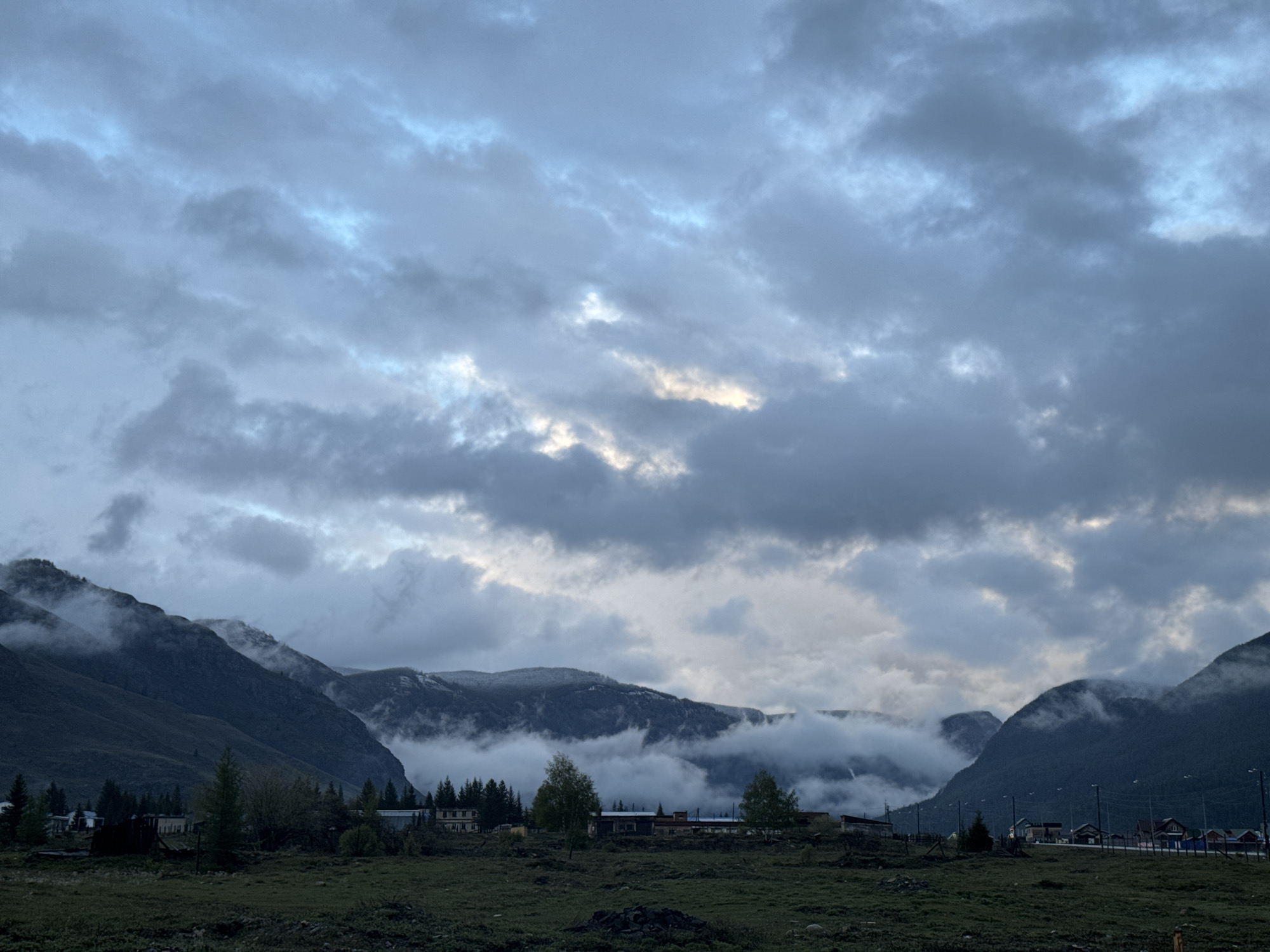 The image shows a landscape in the city of Aktash, Russia. The foreground is dominated by a grassy field. Buildings are visible amidst scattered trees in the middle ground. Beyond this, the scene is defined by mountain ranges partially obscured by low-hanging clouds or fog. The sky is heavily overcast with a mix of grey and blue clouds. The overall lighting suggests a dim or overcast day.
