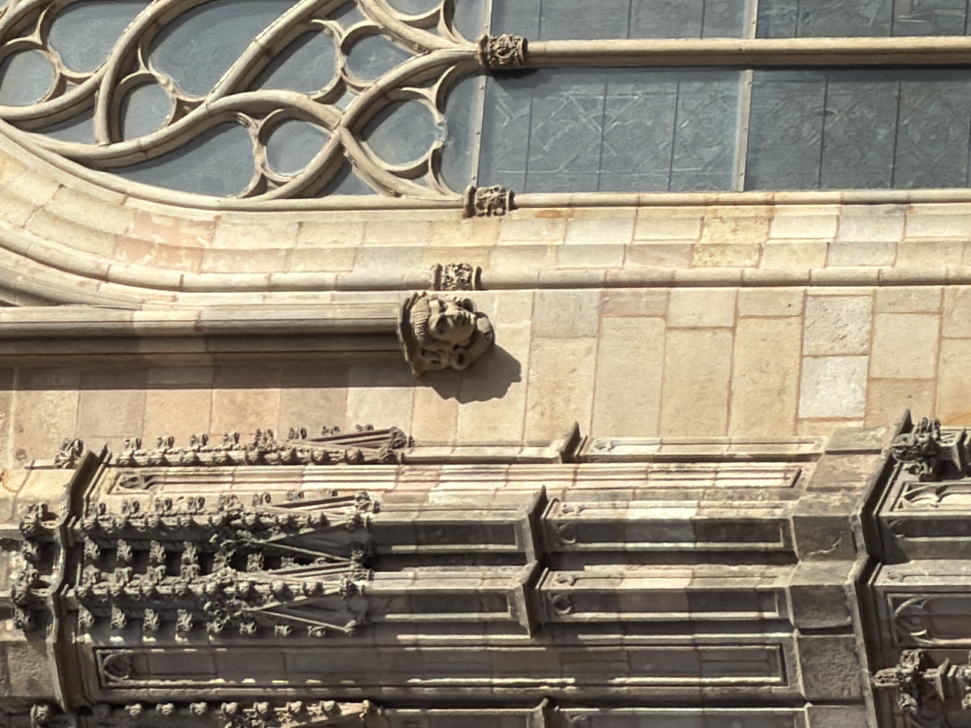 The image shows a close-up detail of the exterior stonework of a Gothic cathedral in Barcelona, Spain. On the left, a section of a buttress or tower is visible, characterized by intricate carvings and vertical fluting. The stone is weathered and appears to be a light beige color. To the right of this, a section of the cathedral wall is constructed of larger, rough-hewn stone blocks. Prominently featured on this wall is a small, carved grotesque or gargoyle, appearing as a face with stylized hair and an open mouth. Further to the right is a section of a large, arched window with tracery, characteristic of Gothic architecture. The window is filled with panes of textured, dark blue or gray glass. The lighting suggests a sunny day, casting shadows and highlighting the textures of the stone. The overall impression is one of ancient, detailed masonry.