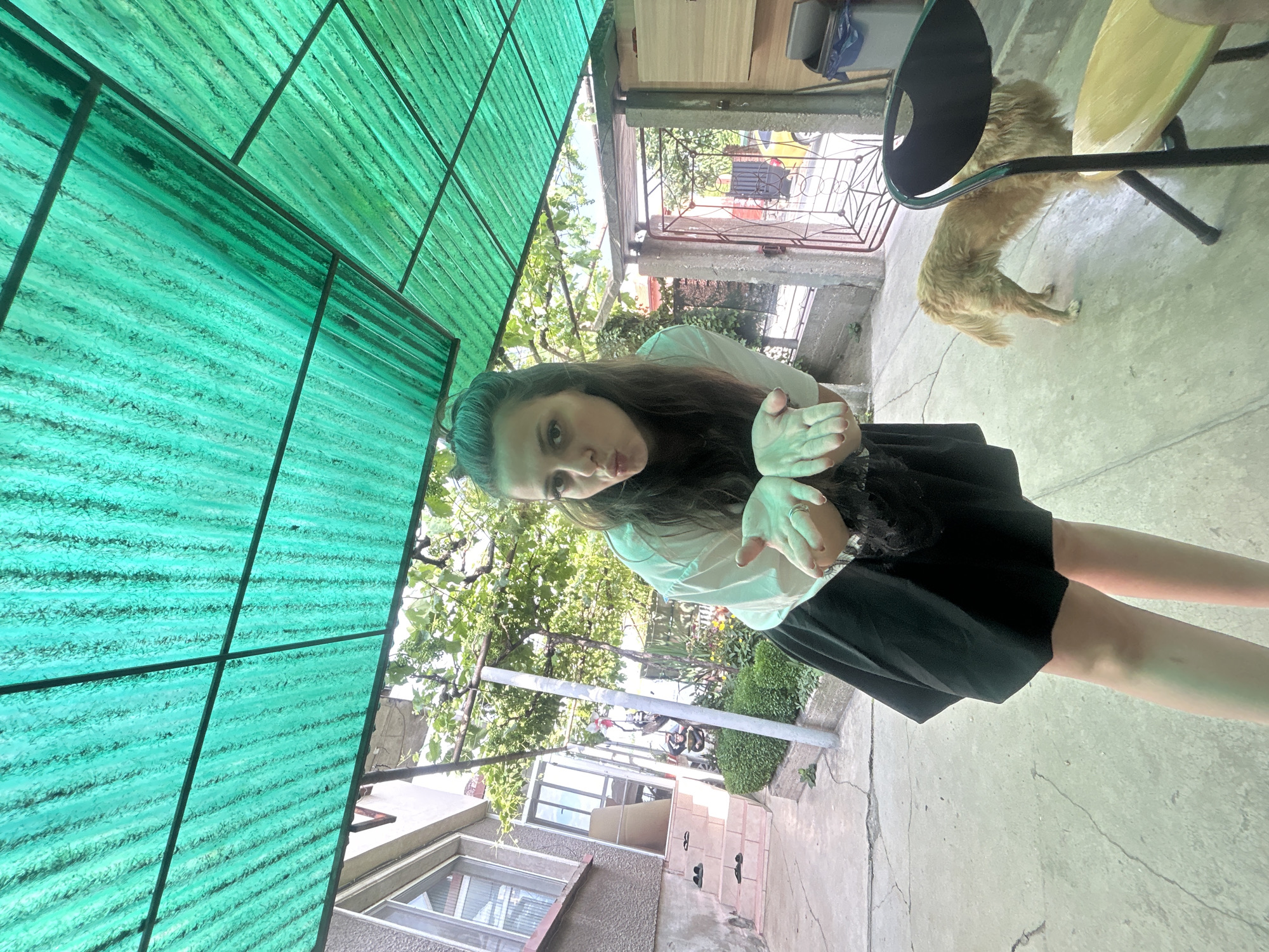 The image depicts a woman with dark hair pulled back, wearing a white t-shirt and a black skirt, posing with her hands extended outwards and her lips pursed. She is standing on a concrete patio in front of a building with a beige facade and a set of stairs leading to an entrance. To the right of the woman, a golden-colored dog is visible, standing and facing away from the camera. The scene is partially covered by a translucent green corrugated plastic roof supported by a metal frame, creating a sheltered outdoor area. In the background, there are trees, vegetation, a gate with a decorative metal pattern, and a yellow vehicle visible through the gate. The location is identified as Govedare, Bulgaria.
