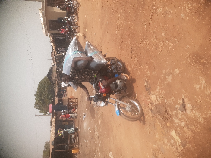 The image depicts an active outdoor market in Gwadanye, Nigeria, under a hazy, light-colored sky. A man, dressed in a dark, patterned short-sleeved shirt and orange flip-flops, operates a red and blue motorcycle (license plate KG A 722 BDG) across an unpaved, reddish-brown dirt road. He transports two large, light-colored sacks, stacked one above the other, on the rear of the motorcycle. In the background, a bustling market environment features numerous individuals, including a woman in a patterned dress near a stall containing yellow containers. A series of makeshift market stalls, constructed from wood and corrugated metal, and simple concrete buildings line the dirt road. A large green tree is visible in the upper left background. Several other motorcycles are parked on the right side of the road, and a small, light-brown dog stands in the mid-ground. The ground exhibits patches of lighter debris and dry earth.