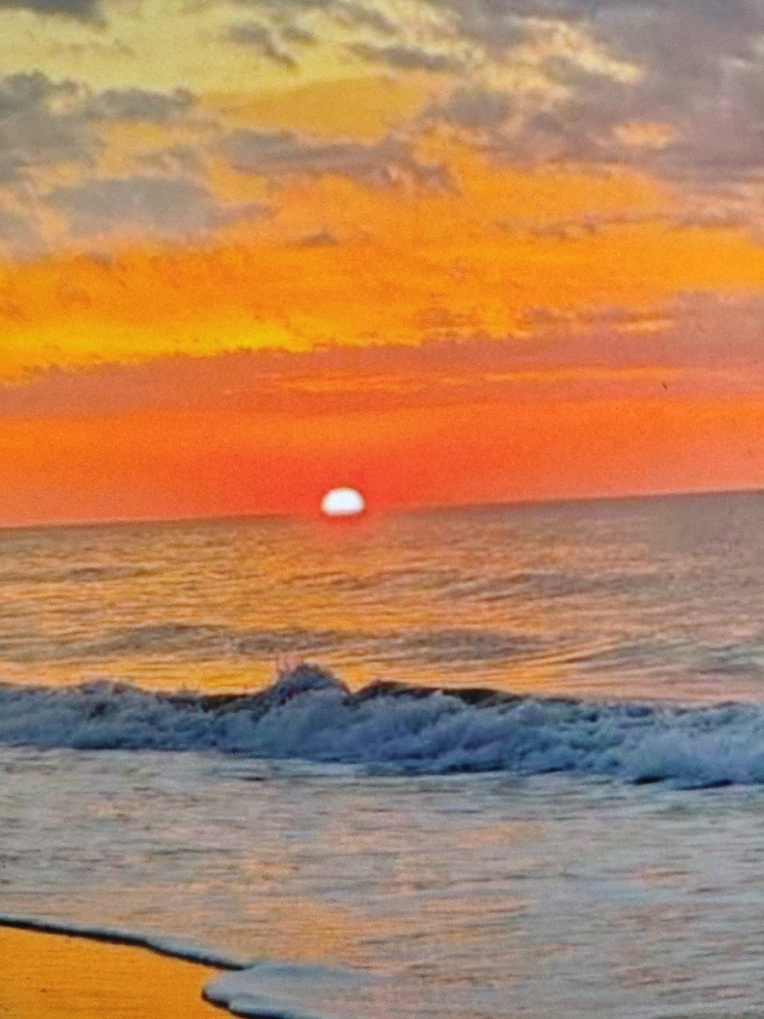 This image depicts a tranquil ocean scene at sunset, with the sun a bright, white orb just above the horizon. The sky is a vibrant gradient of oranges and yellows, with wispy clouds catching the last rays of sunlight. Gentle waves roll towards a sandy shore, which is partially visible in the foreground, glistening with sea foam. The overall impression is one of peaceful natural beauty. There is no indication of the city of Armavir, Russia, in this image, as it depicts a coastal landscape.