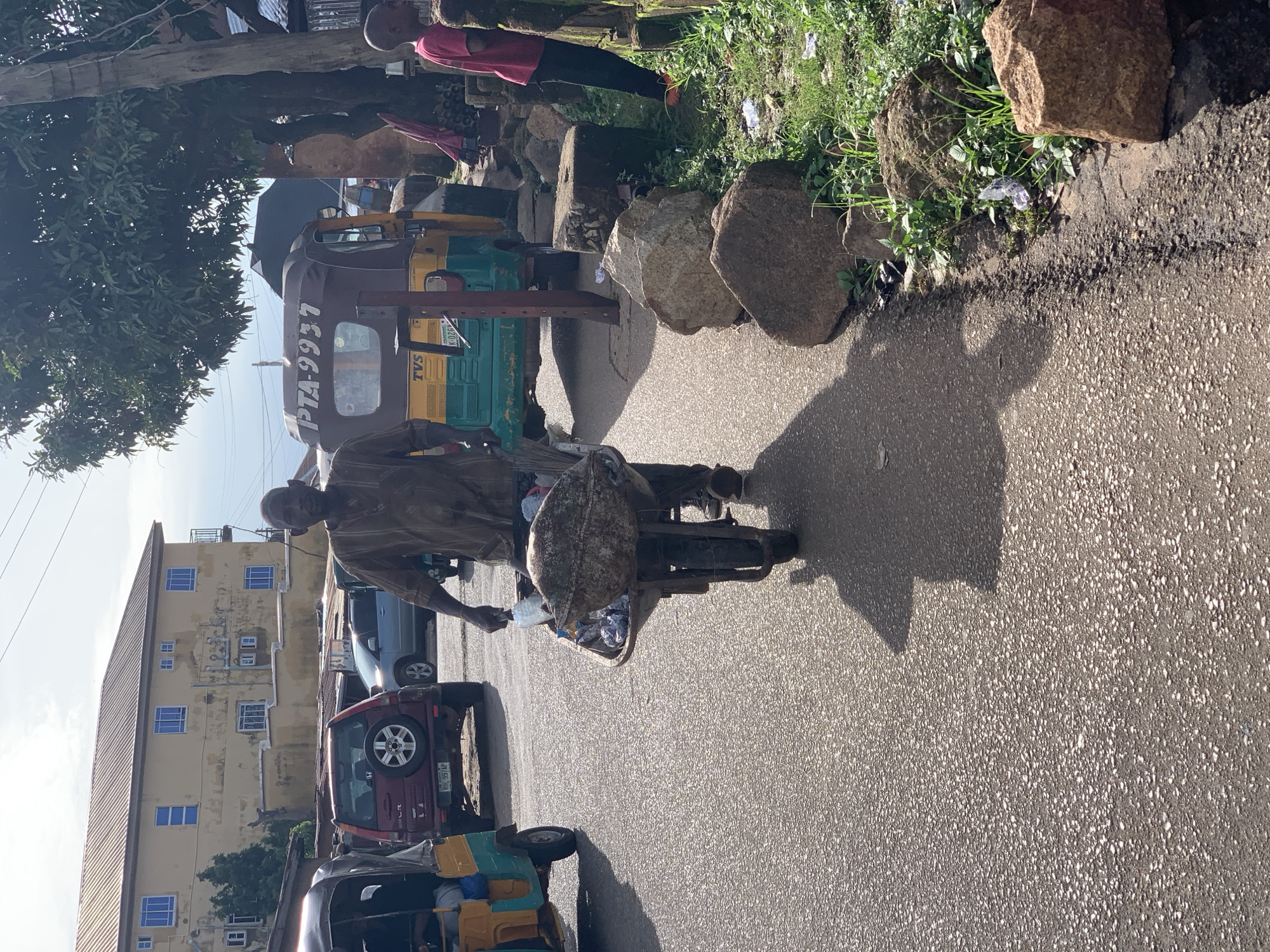 A man is pushing a wheelbarrow full of debris down a paved street. He is wearing a long-sleeved brown tunic. The wheelbarrow contains a large, dark, rounded object, possibly a large piece of refuse or a discarded item, along with other smaller pieces of refuse. In the background, there is a yellow building with blue windows, and a maroon SUV. To the right of the man, a yellow and green auto-rickshaw is parked. Further down the street, a young boy in a red shirt and dark pants stands near a wall. The street is lined with rocks and overgrown green vegetation on the right side. The sky is overcast with some sunlight filtering through. The scene appears to be in Gura Top, Nigeria, a location consistent with the presence of auto-rickshaws and the architectural style of the building.