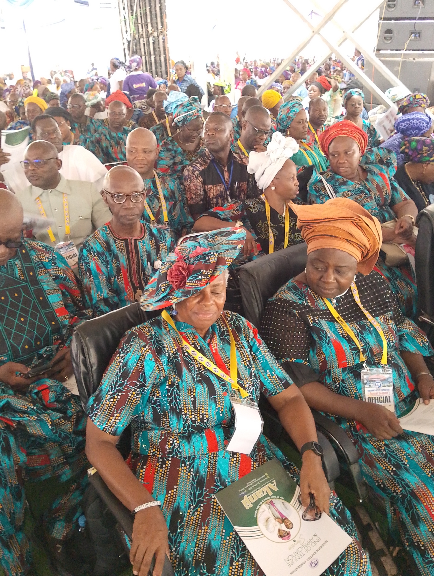 A large group of people, primarily adults of African descent, are seated in rows within what appears to be a spacious tent or canopy structure. The setting suggests an indoor event, brightly lit, likely during the daytime. Most attendees are dressed in vibrant, patterned traditional African attire, featuring shades of teal, blue, brown, red, and yellow. Many women wear elaborate head wraps or decorative hats, while men wear matching tunics or shirts.
The individuals are seated on black cushioned chairs with armrests, indicating an organized gathering. Many wear yellow lanyards with identification badges around their necks. The visible text on some badges reads "OFFICIAL" and "NIGERIAN BAPTIST CONVENTION Annual Session," suggesting a formal convention or conference.
In the foreground, a woman wearing a blue, red, and brown patterned dress and an elaborate matching hat with a floral embellishment smiles subtly. She holds a program booklet titled "Akanji" with further text "NIGERIAN BAPTIST CONVENTION END OF TENURE & APPRECIATION Service." Another woman next to her, wearing a similar dress and a prominent orange headwrap, appears to be observing the event. A man in glasses wearing a light green shirt and an "OFFICIAL" badge is visible in the middle ground, while others in matching patterned clothes are engrossed in the proceedings. The overall atmosphere is composed and attentive, with people mostly facing forward, implying a speaker or stage is present beyond the frame. Some individuals are seen wearing watches, and one is interacting with a mobile phone. The diverse yet coordinated attire creates a visually rich and culturally distinct scene.