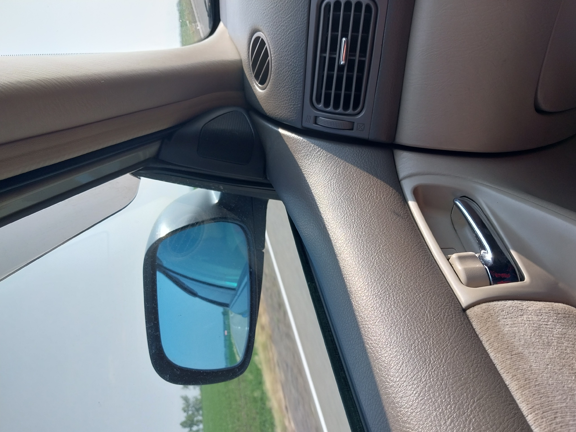 The image is a close-up interior view of a car, taken from the passenger seat looking forward and to the left. A blue-tinted side mirror reflects the road ahead, bordered by green fields. The car's dashboard is visible on the right, featuring air vents and a chrome-accented door handle in the foreground. The sky is overcast and light. The image does not show the city of Sychevka, Russia.
