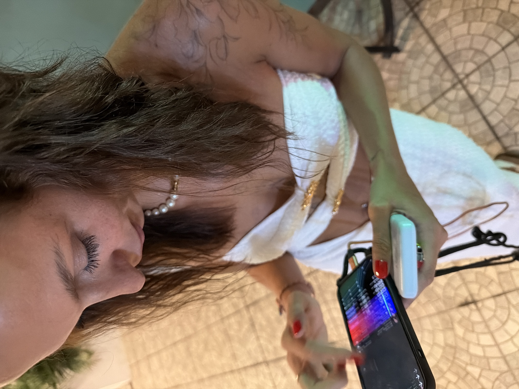 A woman with long brown hair, wearing a white strapless top, stands outdoors on a tiled floor. She is looking down at a smartphone in her left hand, which displays a colorful screen. Her right hand holds a white portable charger. The woman has red nail polish on her fingernails and a pearl necklace around her neck. A tattoo is visible on her left shoulder. The background is blurred, but appears to be an outdoor setting with some greenery and a chair. The scene is consistent with being in a city like Ban Rang Rak, Thailand.