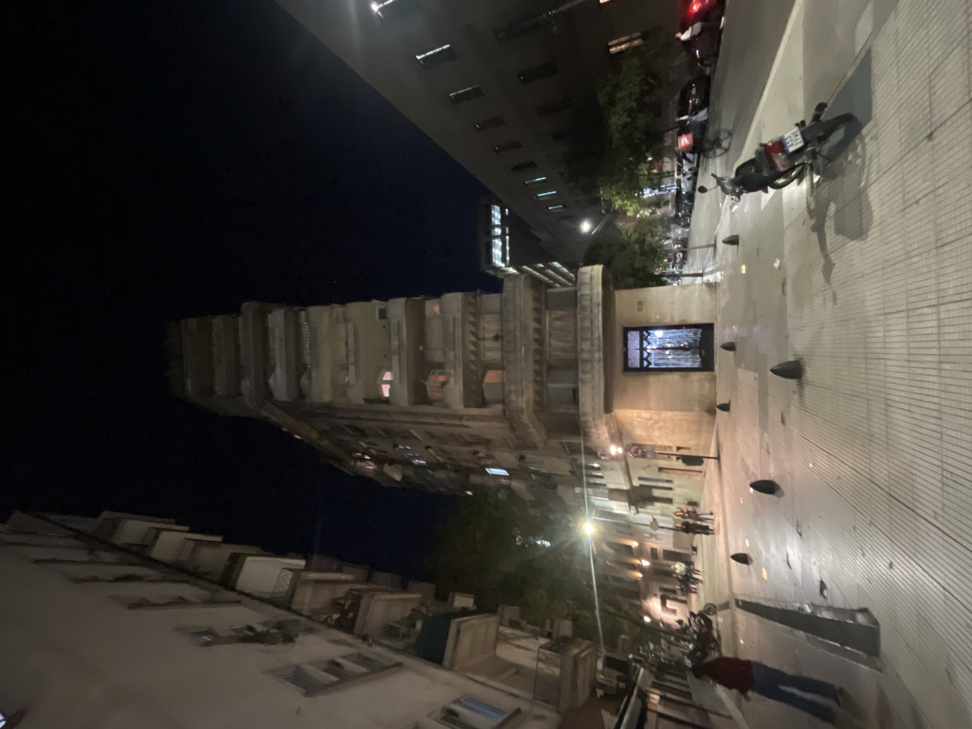 The image shows a street scene in Buenos Aires, Argentina at night. On the right, a tiled sidewalk with conical bollards lines the street. Several motorcycles are parked along the sidewalk. People walk along the street in the distance. The Kavanagh Building, a notable skyscraper in Buenos Aires, is visible on the left side of the frame. It is a tall, stepped building with visible windows and architectural details. Streetlights illuminate the scene. Other buildings with windows are visible at the top and bottom of the frame. A person is walking with a red shirt.