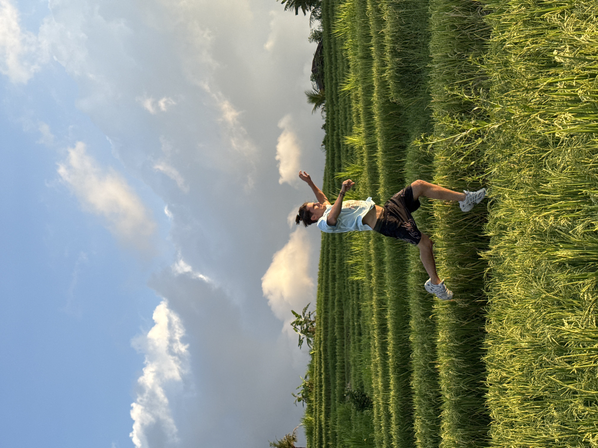 A man in a light blue shirt and black shorts is standing in a lush green rice paddy in Padangbulia, Indonesia. He has his arms raised, seemingly mid-motion. The rice paddy is tiered, creating a stepped landscape. The sky overhead is blue with scattered clouds, some of which are dark.