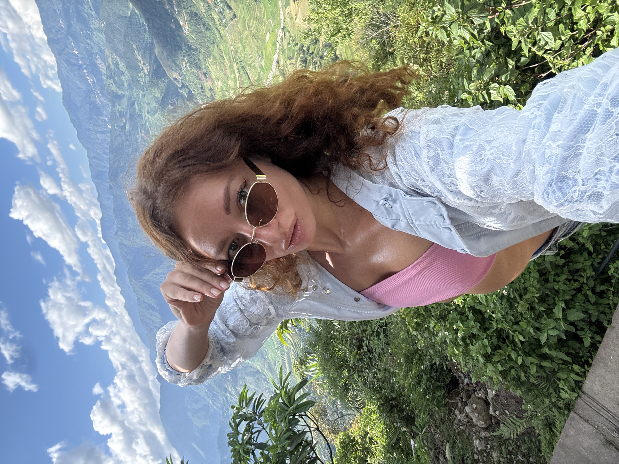 A young woman with long, curly reddish-brown hair is centered in a selfie, looking directly at the camera with slightly parted lips. She wears oval, brown-tinted sunglasses with a thin gold-colored frame, a pink ribbed crop top, and a light-colored jacket featuring a visible lace overlay pattern. Her right hand is raised, with fingers near her temple and the sunglasses frame. The background presents a panoramic view of a vast mountainous landscape under a bright blue sky with scattered white clouds. Multiple layers of mountains, covered in dense green vegetation, recede into the distance. Extensive terraced fields, characteristic of rice paddies, are visible on the mountain slopes and in the valleys, alongside scattered small structures or settlements. The immediate foreground on the left includes green foliage, a plant with broad leaves, and a concrete edge. The scene is well-lit by natural sunlight, consistent with the geography of Tu Le, Vietnam.