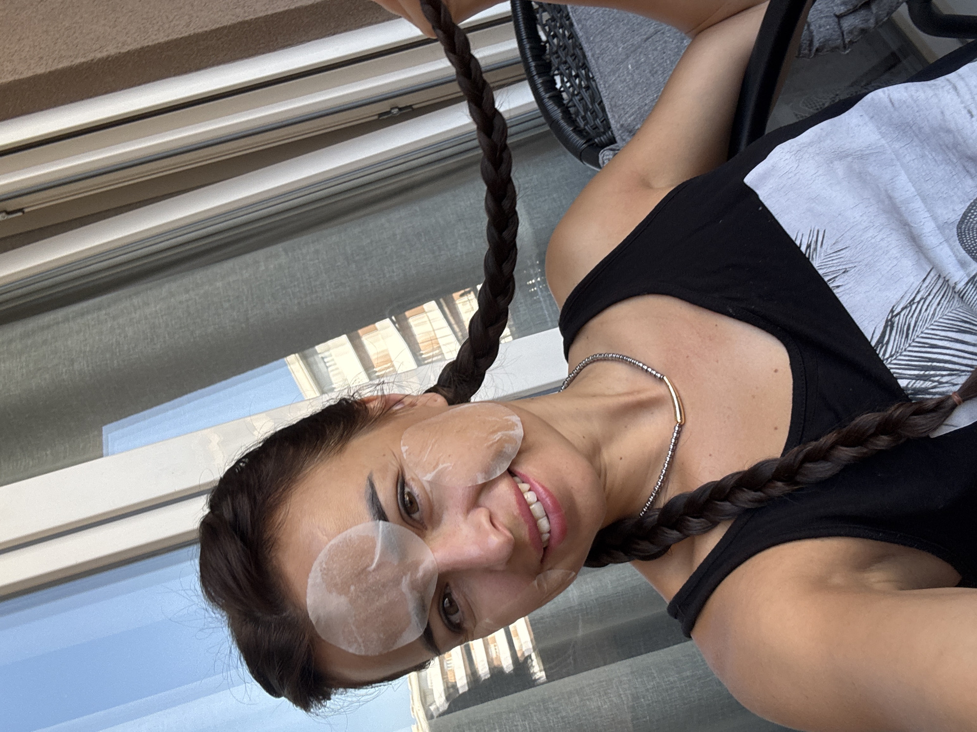 A medium shot shows a woman with dark hair in two braids. She is wearing a black tank top and a necklace. She is smiling and has clear patches on her face, likely for skincare. The background consists of a window with blinds and a balcony railing. The lighting suggests it is daytime.