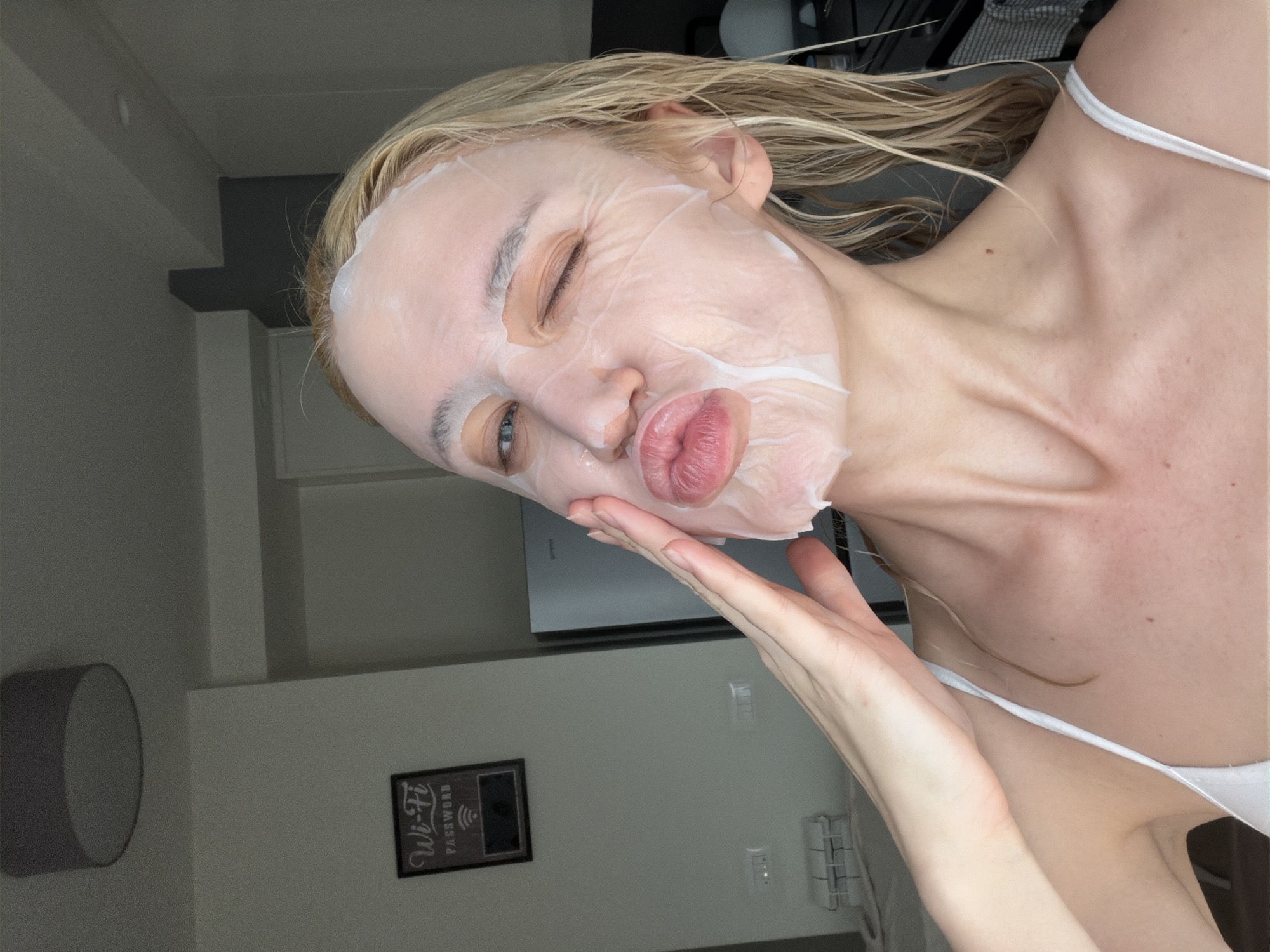 A light-skinned woman with wet, blonde hair is captured in a close-up, angled shot, likely taking a selfie indoors in what appears to be a residence or hotel in Belgrade, Serbia. She is wearing a white sheet face mask that covers most of her face, with cutouts for her eyes, nose, and mouth. Her left eye is open and appears to be winking, while her right eye is closed. Her lips are playfully pursed in a pout, highlighted by vibrant pink lipstick. Her left hand is raised to her chin, gently cupping her jawline, contributing to the posed nature of the image.
She is dressed in a simple white spaghetti-strap top. Her neck and prominent collarbone are visible, showing clear skin with a small mole on her left collarbone. Her wet hair suggests she might have recently washed it or is refreshing herself.
The background features a plain, light grey wall. On the left side, a round, wall-mounted light fixture is partially visible. Towards the bottom right, a framed black sign hangs on the wall, explicitly displaying the words "Wi-Fi" and "PASSWORD" in white text. Further in the background, a large, light-colored appliance, possibly an air conditioner or refrigerator, can be vaguely discerned. The overall ambiance suggests a relaxed moment of personal care, conveyed with a playful and lighthearted mood.