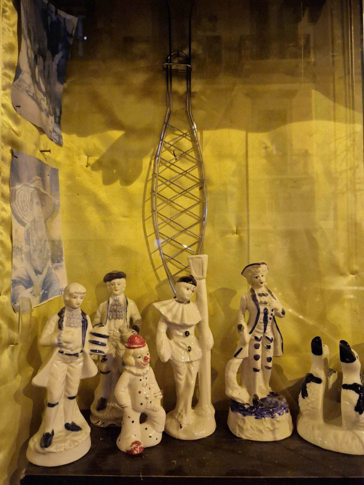 The image shows a collection of ceramic figurines displayed on a shelf against a textured yellow background. The figurines include several figures in historical attire, a clown with a red hat and polka dots, and two black and white dog figurines. A metal object, possibly a decorative piece or a tool, hangs from the top of the frame. To the left of the figurines, two black and white prints are pinned to the background. The scene appears to be in a display case or on a shelf in a home or shop. The provided context states that the image was taken in Taranto, Italy.