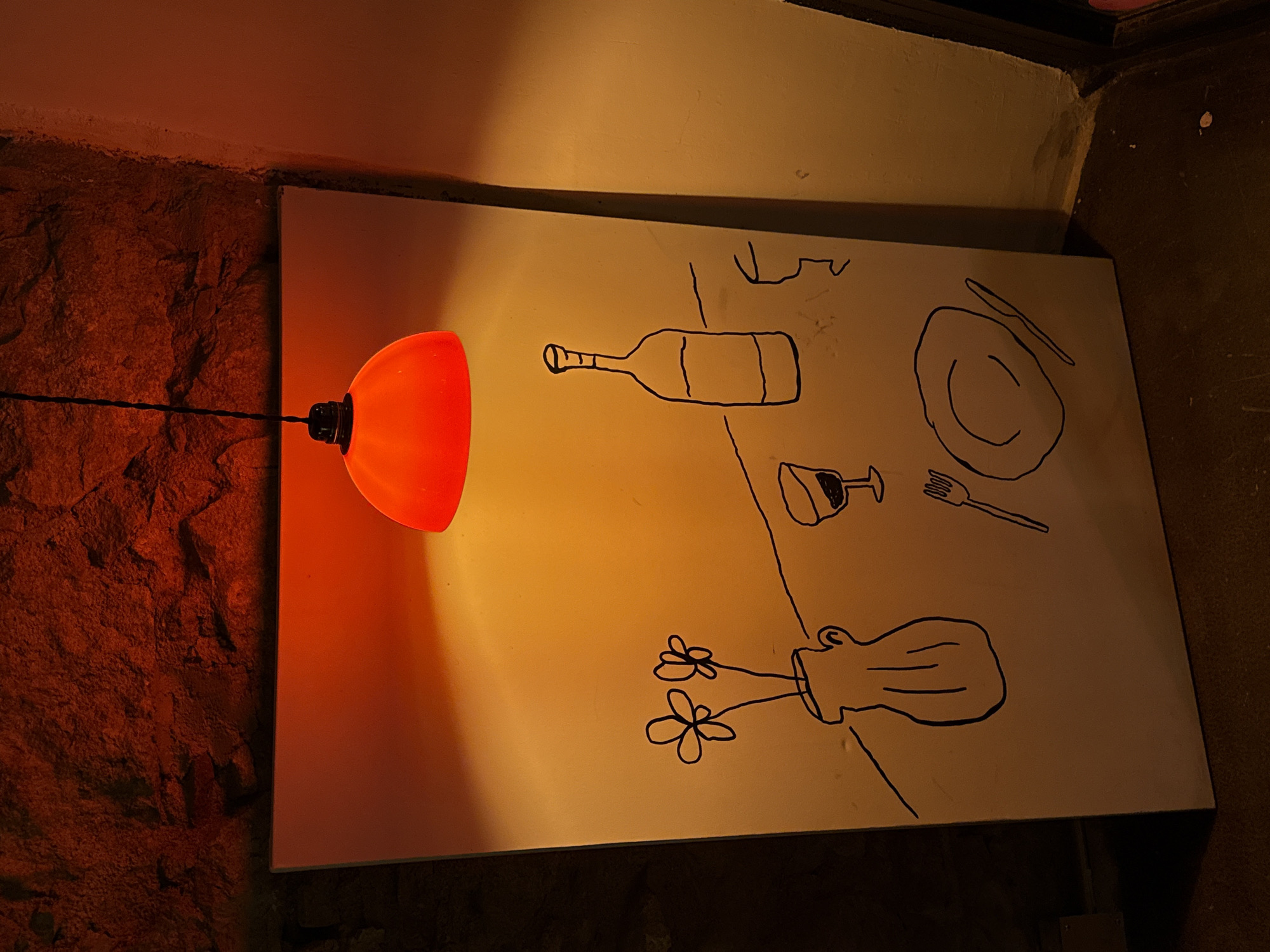 The image depicts a drawing illuminated by an orange light. The drawing is a simple, childlike illustration of a wine bottle, glass, plate with silverware, a vase, and flowers. The drawing is on a rectangular white surface, likely a canvas or board, positioned near a textured, reddish-brown stone or brick wall on the left. An orange lamp is attached to the wall adjacent to the drawing and projects its light onto the artwork. The scene has dim lighting overall. Based on the subject matter depicted in the drawing (wine, food) and the ambiance, it's likely the scene is in a restaurant or bar. However, without additional contextual information, it's impossible to confirm it's located specifically in Barcelona, Spain.
