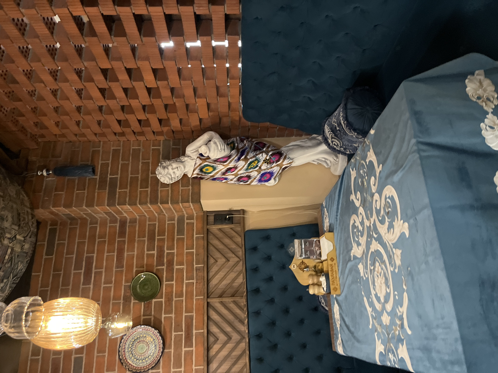 This image captures the interior of what appears to be a restaurant or cafe in Izhevsk, Russia, showcasing an eclectic and richly decorated dining space. The perspective is from a slightly elevated, angled position, looking across the room.
The decor features a blend of rustic, classical, and vibrant elements. Walls are constructed from exposed red brick, and a distinctive ceiling is made of interlocking brick-like pieces, forming an open lattice pattern. Running along one side is plush, dark teal upholstered banquette seating with a button-tufted design.
A prominent feature is a white, classical-style reclining statue, possibly depicting a mythological figure, placed on a ledge against a brick wall. It is adorned with a colorful fabric featuring intricate, geometric, and eye-like patterns, resembling Ikat textiles. A round, dark blue decorative cushion rests on the banquette next to the statue.
In the foreground, a table is covered with a light blue tablecloth detailed with elegant white scroll and floral patterns. On the table, a wooden stand holds small, lidded containers, likely for condiments or sugar. A small box also sits on the table, bearing the Cyrillic text "ПАХЛАВА" (Pakhlava) and "ЛЮБИМАЯ" (Lyubimaya), indicating a sweet pastry product like baklava.
Other details include a large, hanging glass light fixture emitting a warm, golden glow, and two decorative plates mounted on the brick wall. A section of wall near the table features wooden panels arranged in a chevron pattern, adding another textural dimension to the space. The overall ambiance is warm and inviting, created by the interplay of materials, colors, and lighting. No people or specific activities are visible, emphasizing the static beauty of the interior design.
