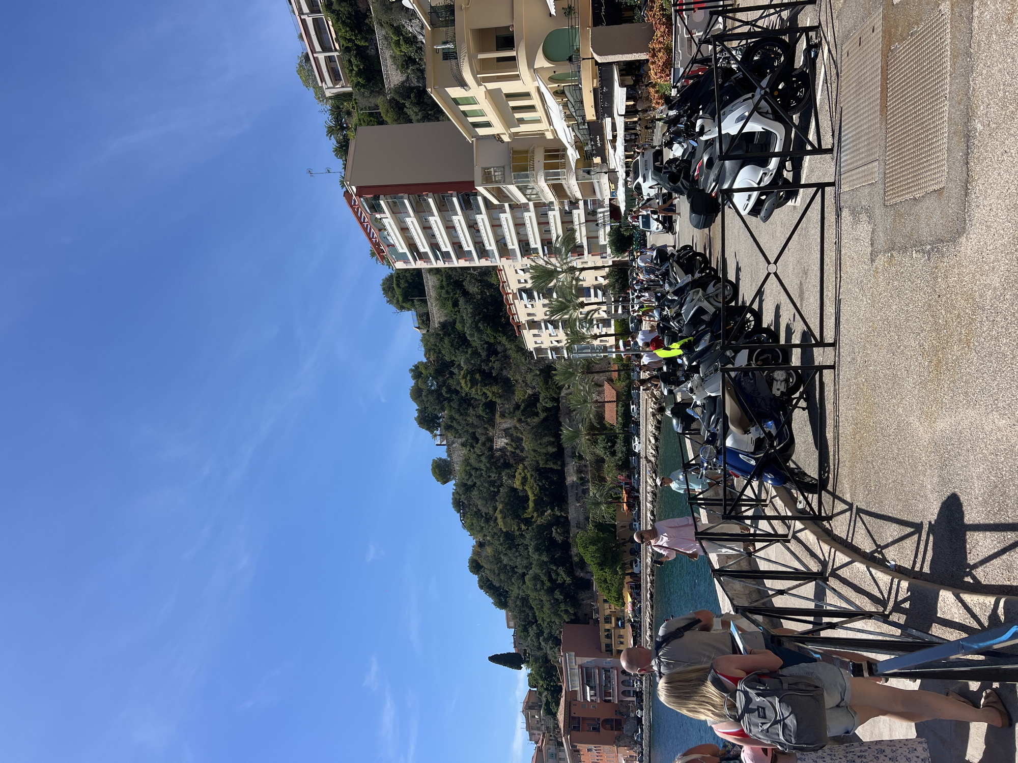 This is a medium shot, eye-level, outdoor, daytime, vertical photograph of a waterfront promenade in Villefranche-sur-Mer, France. The promenade is paved with concrete and lined with a black metal fence. Behind the fence, numerous parked motorcycles and scooters are visible. Beyond the vehicles, a street runs parallel to the promenade, with cars and more motorcycles parked along it. Several buildings of varying architectural styles and heights are situated on the hillside overlooking the street, including a tall, modern, multi-story building with balconies and red accents, and a cluster of older, ochre-colored buildings. Lush green trees and vegetation cover the hillside. To the left of the promenade is a body of blue water, likely the Mediterranean Sea, with a few boats visible in the distance. In the foreground, on the promenade, four people are visible. Closest to the camera is a woman from the back, wearing a grey backpack, denim shorts, and a red top. To her left is another woman, partially visible, wearing a patterned dress. A man with a grey backpack is standing to the right of the first woman, facing the water. Further back, a woman in a pink top and headscarf is standing and holding a phone, appearing to take a picture. A man in a light blue shirt is walking towards the water. The sky is a clear, bright blue with a few wispy white clouds.