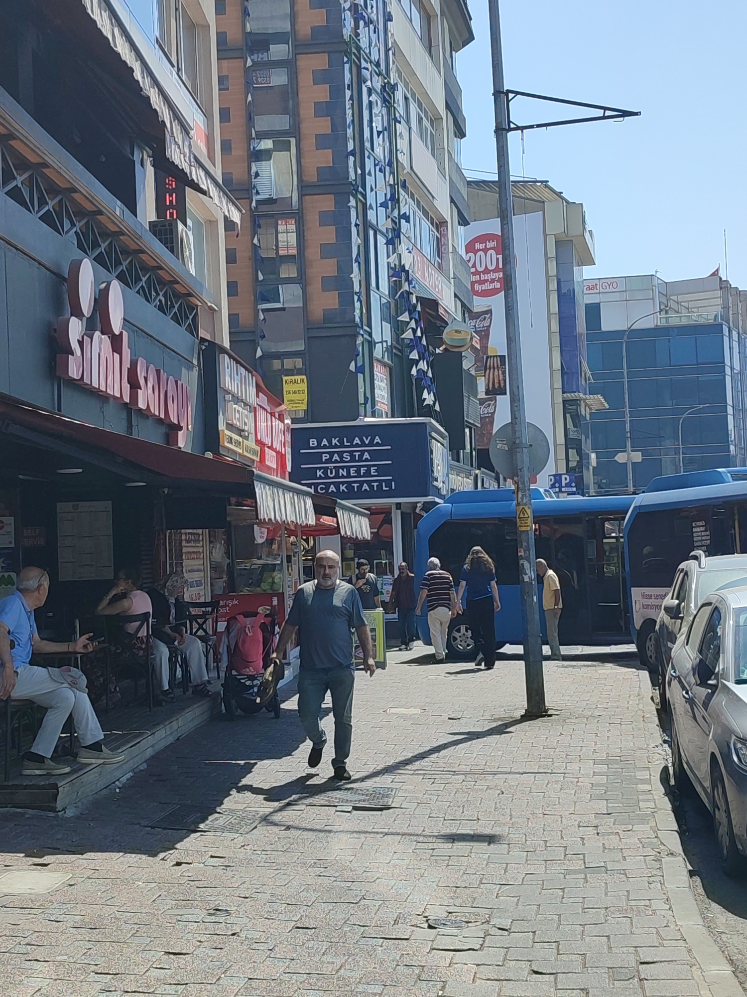 The image depicts a bustling street scene in Kadıköy, Turkey, on a sunny day. In the foreground, a man is walking away from the camera on a cobblestone sidewalk. He is wearing a dark t-shirt and jeans. To his left, several people are seated at tables outside a shop with a red awning. The shop appears to be a bakery or cafe, with signage visible including "Simit Sarayı."
Behind the seated individuals, a blue bus is parked, and people are boarding or disembarking. More people are walking along the sidewalk and the street. The background features a multi-story building with modern glass architecture and some older buildings with visible signs in Turkish. Flags are also seen hanging from some buildings. The overall environment suggests a lively urban commercial area.