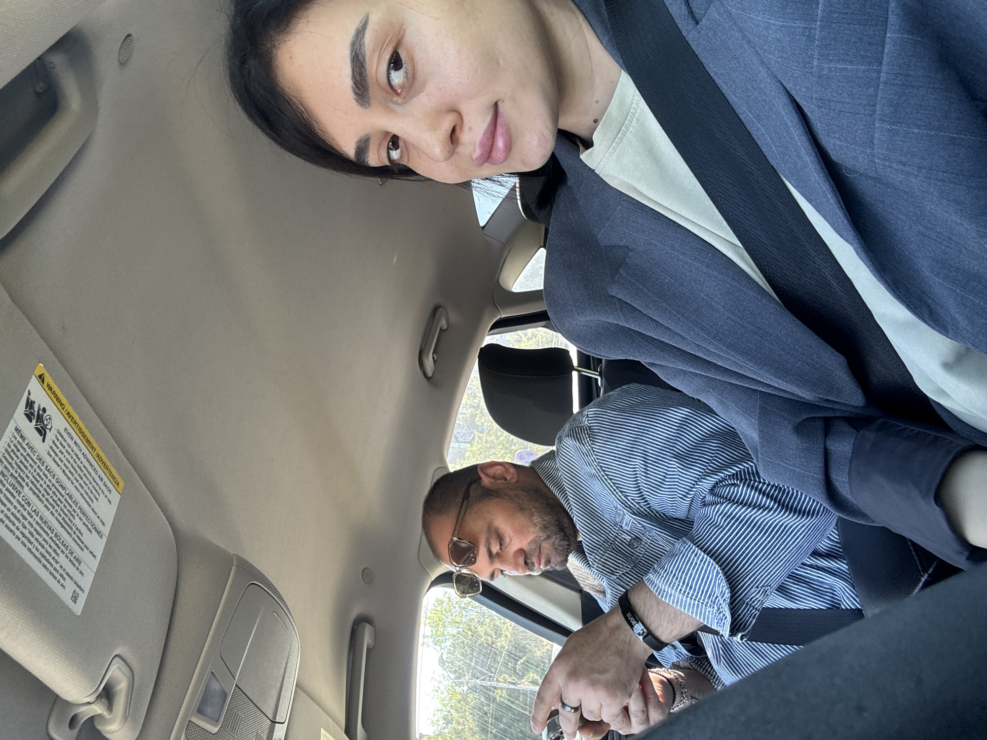 The image shows two people inside a car. A woman with dark hair is positioned higher in the frame, appearing to be taking a selfie. She is wearing a gray blazer. A man with glasses and a striped shirt is visible below, seemingly in the driver's seat. The scene takes place inside the car, with the car's interior including the dashboard, sun visor with warning label, and seat visible. Outside the car window, trees and daylight indicate an outdoor environment. It is not possible to determine from this image the exact location in Tbilisi, Georgia.