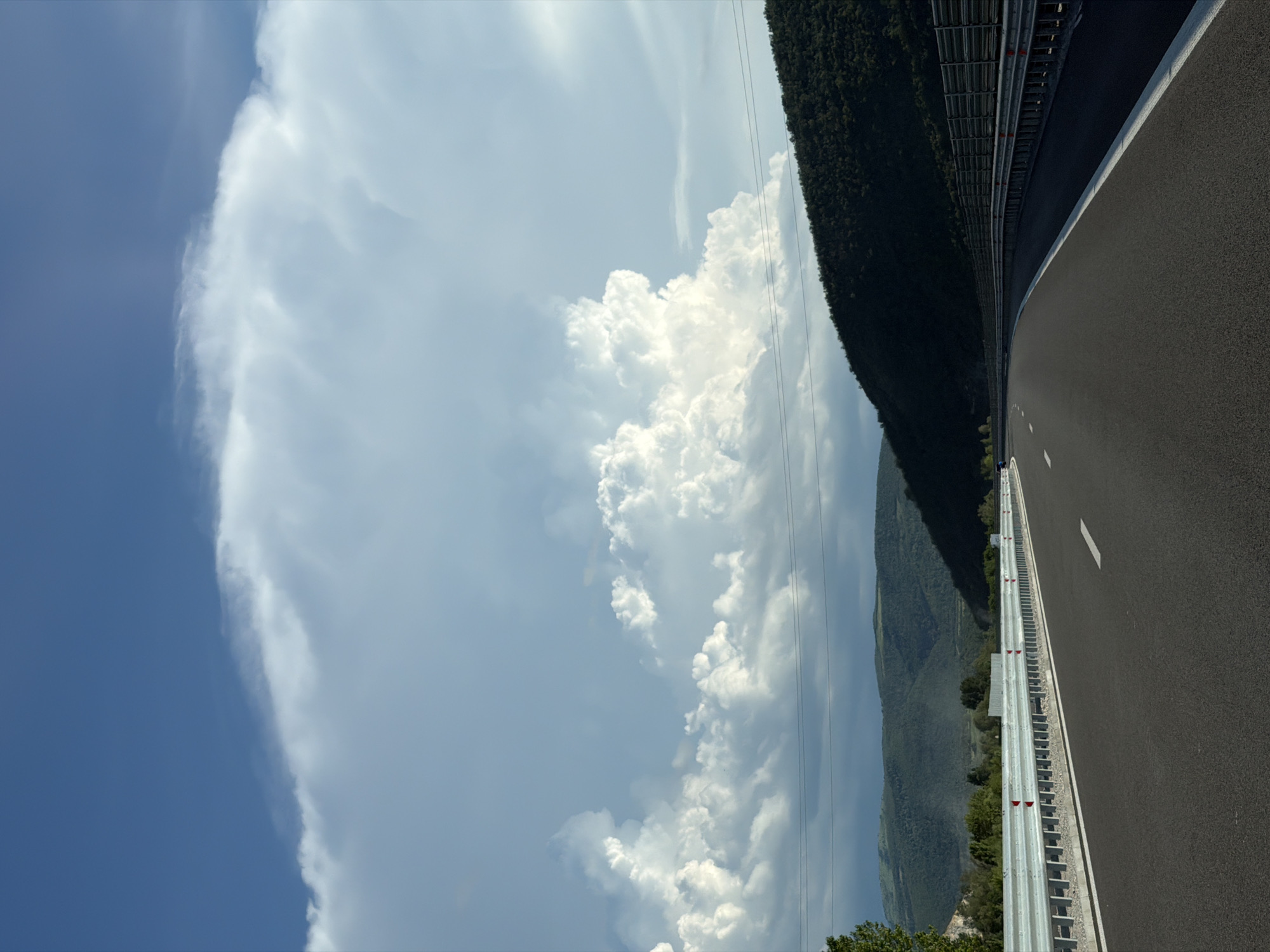 The image shows a highway with a dark asphalt surface and white lane markings. On the left side of the road, there is a metal guardrail with red and white reflectors. Beyond the guardrail, there are trees and hills covered in vegetation. On the right side of the road, there is another metal guardrail, and further in the distance, a dark, densely forested hillside slopes upwards. The sky is a vibrant blue with large, dramatic white cumulus clouds. The road appears to be in a rural or semi-rural setting, possibly on the outskirts of Peshterna, Bulgaria, given the mountainous terrain and vegetation. There are no people or vehicles visible in the immediate scene. The lighting suggests it is daytime, with the sun likely to the right, casting shadows on the hillside. Power lines are visible strung across the landscape.