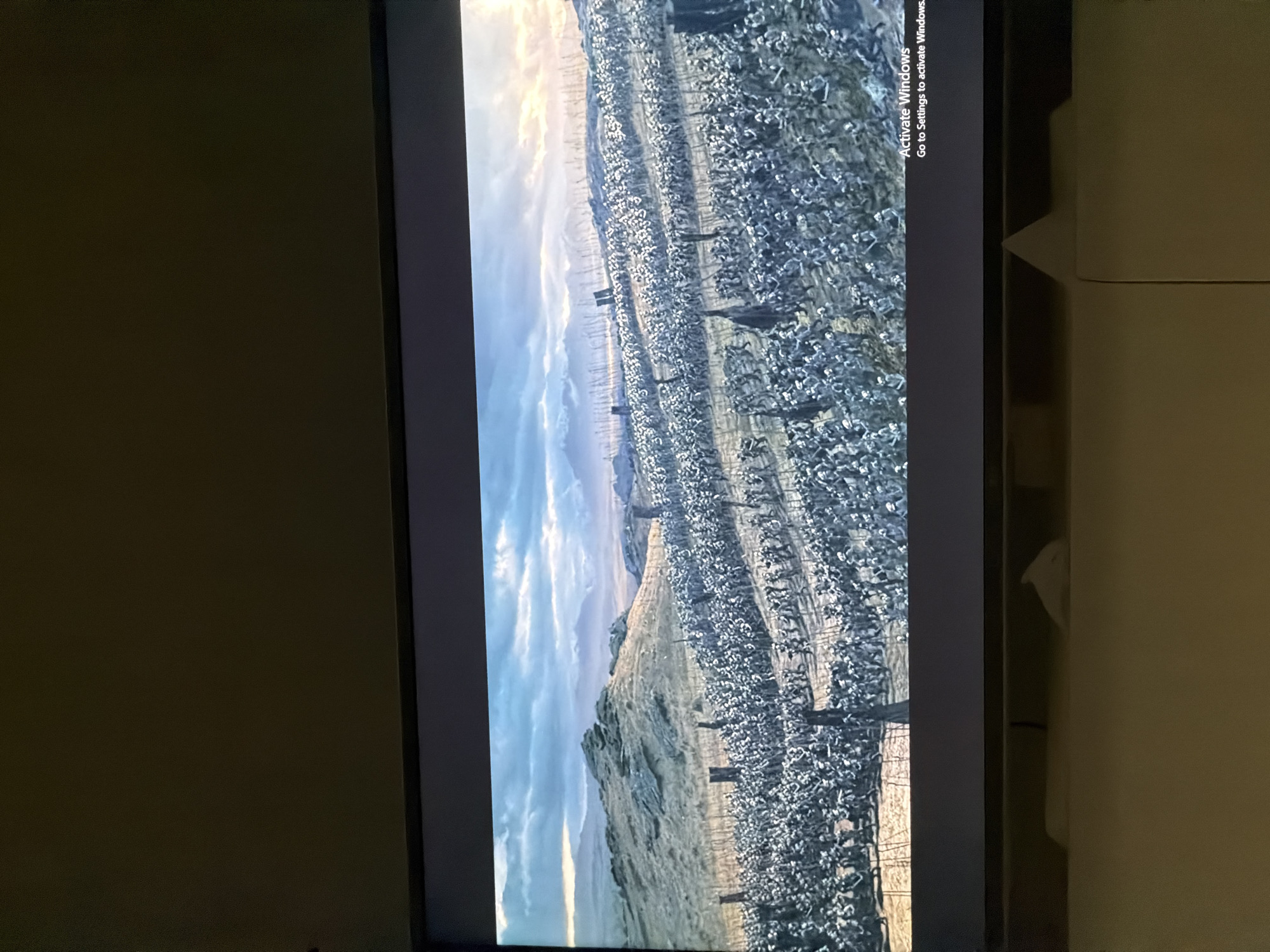 The image captures a horizontal screen displaying a vast, panoramic landscape, likely from a video game or CGI scene, set in an indoor environment in Plovdiv, Bulgaria.
**Subjects:**
On the screen, a sprawling field of countless small, white, textured objects dominates the foreground and midground. These objects are arranged in distinct rows and could represent cotton plants, stylized skeletons, or a massive army formation. Dark, vertical structures, possibly trees or pillars, are scattered throughout this field. In the background, rolling hills and distant mountains are visible beneath a partially cloudy sky. Towards the far left of the screen, numerous tall, slender structures, resembling wind turbines or communication towers, extend into the horizon.
Off-screen, the immediate surroundings indicate an indoor setting. To the left and right of the screen, dark, muted walls or furniture are visible. On the right side, a light-colored object, possibly a folded piece of clothing or a towel, can be seen hanging on what appears to be a shelf or inside a cabinet.
**Activity or event taking place:**
The screen is actively displaying a still image or a paused scene, presenting a large-scale, detailed environment. The primary activity is a photograph being taken of this illuminated display.
**Notable details:**
The time of day depicted on the screen appears to be either sunrise or sunset, characterized by soft, warm light near the horizon and cooler blue tones in the upper sky. The weather in the displayed scene is cloudy. The landscape on the screen has a unique, possibly stylized or fantastical quality due to the unusual ground cover and distant structures.
The location of the photograph itself is Plovdiv, Bulgaria.
**Visible text:**
In the bottom right corner of the displayed screen, the text "Activate Windows" and "Go to Settings to activate Windows." is clearly visible, indicating that the operating system running on the device is not activated.