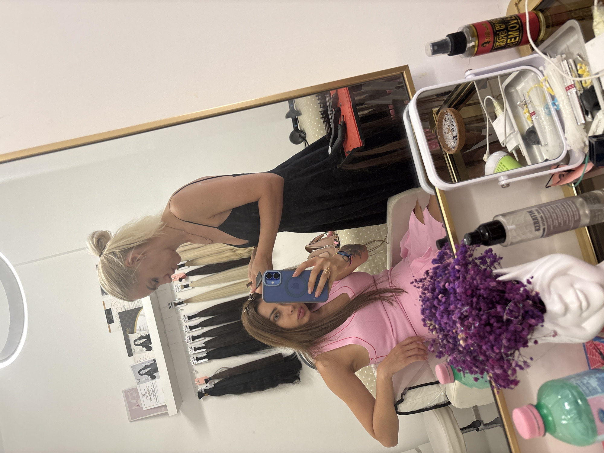 A woman with blonde hair tied in a bun, wearing a black slip dress, styles the hair of another woman in a pink dress in a salon. The woman in the pink dress is sitting in a chair and holding up a blue smartphone, taking a mirror selfie. The salon appears to be a hair extension salon, with numerous bundles of hair extensions hanging on the wall behind the women. There are also various hair care products and tools visible in the foreground and reflected in a mirror. The scene is well-lit, suggesting an indoor setting. The image does not contain any specific indicators of its location in Rome, Italy.