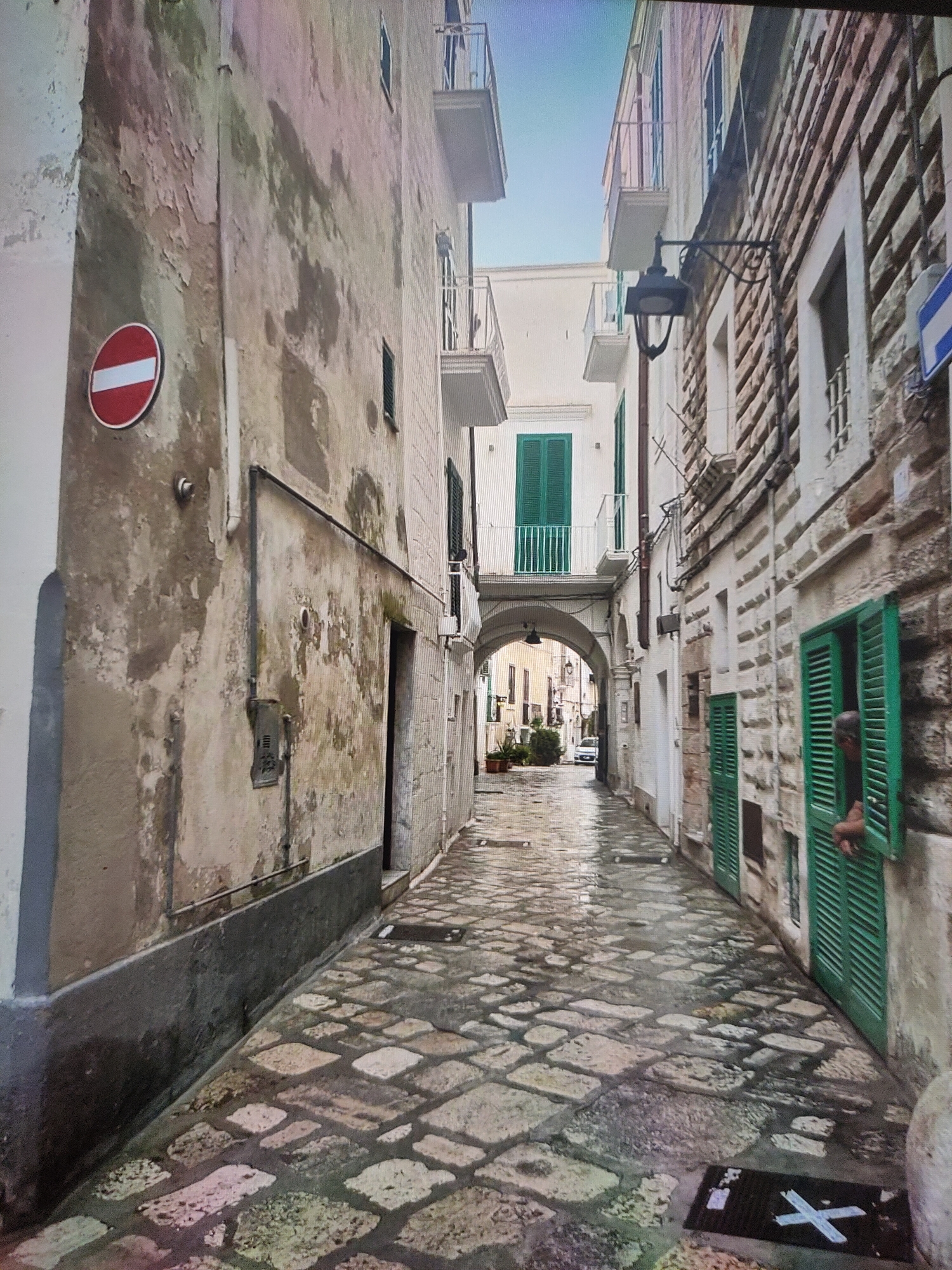 The image depicts a narrow, wet cobblestone alleyway in an old European city. The buildings lining the alley are constructed from stone and plaster, with weathered facades displaying peeling paint. On the left, a building has a red and white circular "no entry" sign. Green shutters are visible on windows and doors of the buildings. An arched passageway is visible further down the alley, leading to an area with more buildings and a car. A person's arm and shoulder are visible leaning out of a green-shuttered window on the right. The sky is a bright, clear blue. The overall impression is of a historic urban environment that appears to have recently experienced rain, given the wet pavement and reflections. This scene is not located in Zrenjanin, Serbia.