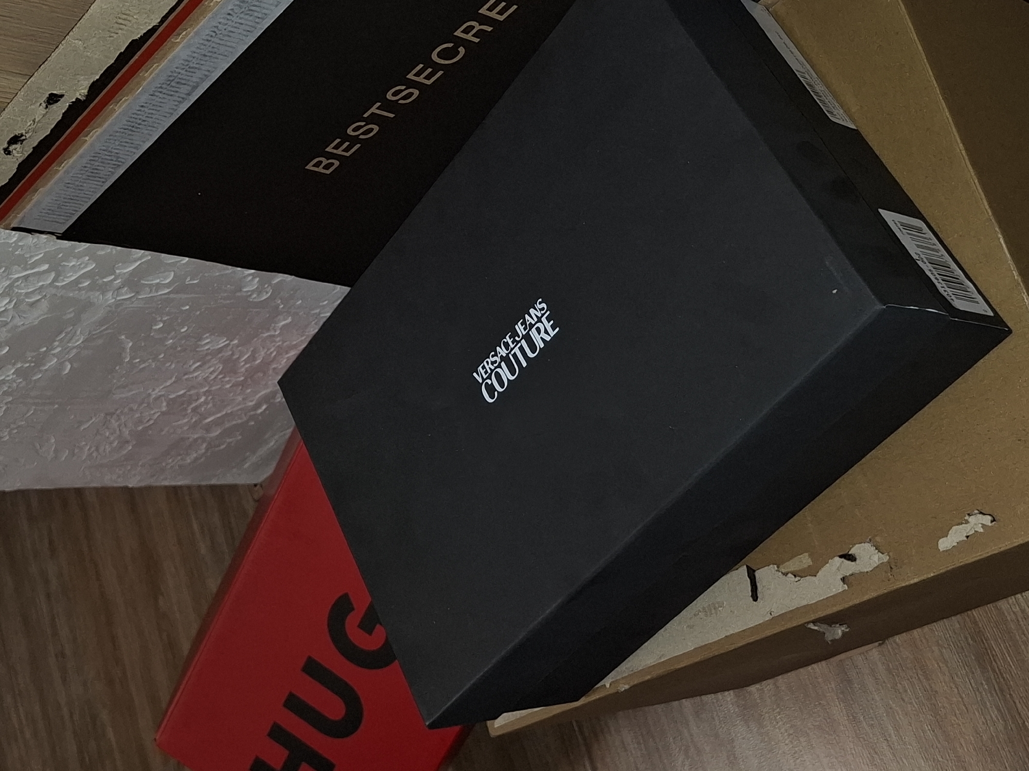 The image shows a stack of cardboard boxes with brand names visible. A black box labeled "VERSACE JEANS COUTURE" sits on top of a red box with the partial word "HUGO" visible. To the left is a dark brown box with the word "BESTSECRE" appearing in gold lettering. A larger, tan cardboard box supports the arrangement on the right. The background is a wall with a textured, possibly painted, surface. The location, Veliki Preslav, Bulgaria, cannot be definitively determined from this image.
