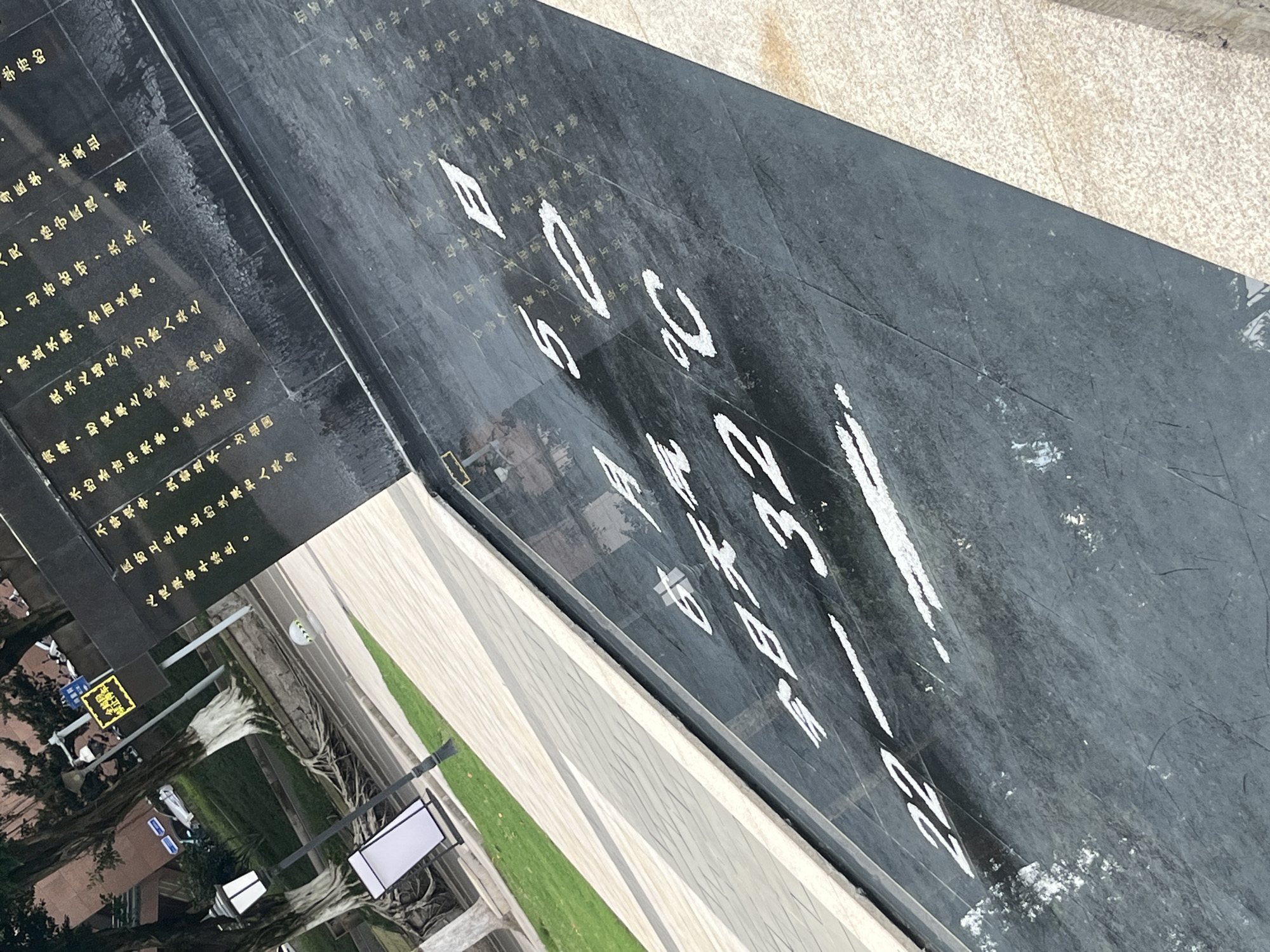 The image presents an outdoor scene in Yuzhong District, China, likely a public space or campus area, under what appears to be an overcast sky during daytime. The central focus is a large, dark, polished stone monument or plaque set into the ground at an angle.
The monument features extensive Chinese characters in a gold or yellow color, which are part of a professional pledge or declaration. OCR analysis reveals phrases emphasizing medical ethics, including "love the motherland," "abide by medical ethics," "do my utmost to eliminate human suffering," "uphold the sanctity and honor of medical skills," "save the dying and heal the wounded," and a lifelong commitment to "fight for the development of the motherland's medical and health cause and for human physical and mental health." This suggests the monument is dedicated to medicine or a related institution.
Additionally, prominent white characters and numbers are displayed on a slightly different section of the monument, clearly showing "22-32℃." Other white letters and numbers, such as "E", "S", "B", "C", "3", and "2" are also partially visible.
The surrounding environment includes a patterned, light-colored paved walkway next to a strip of green grass. Beyond this, a street is visible with trees and a light pole. A yellow street sign in the background displays Chinese characters that translate to "height limit" (限高) and "pay attention to safety" (注意安全). No specific human activity is captured in this static scene, and the monument itself shows some smudges or wear.